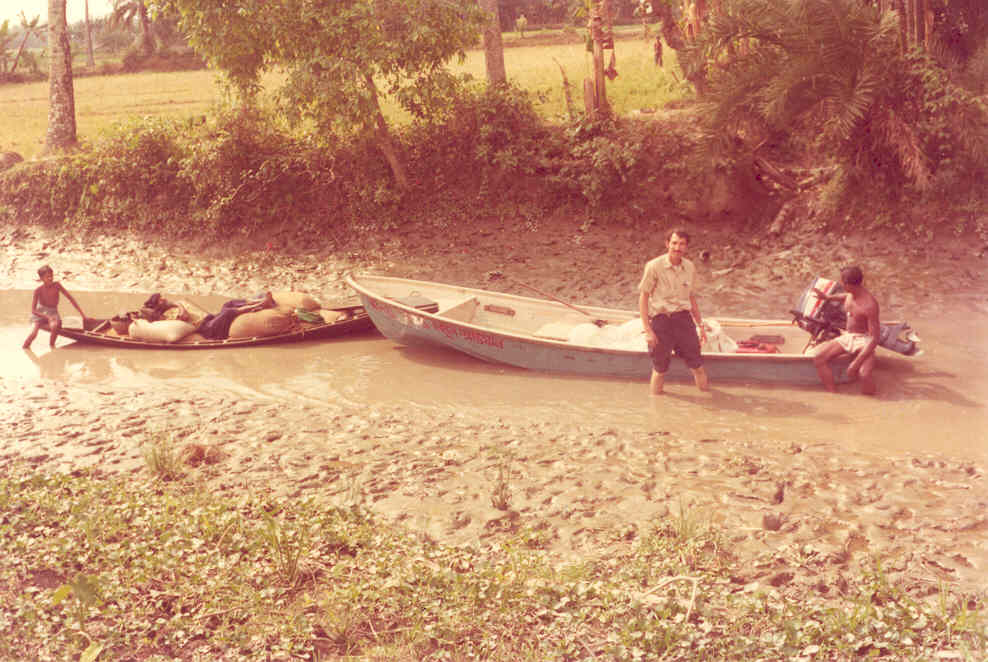 Two boats are in very shallow water and mud. On the right is a motorboat on which a man sits beside the outboard motor, which has been tilted into the boat, and another stands in front of the boat with mud up to his calves. A child and filled sacks are on the smaller boat to the left.