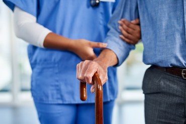 A photo of a nurse helping an older man walk with a cane in a nursing home.