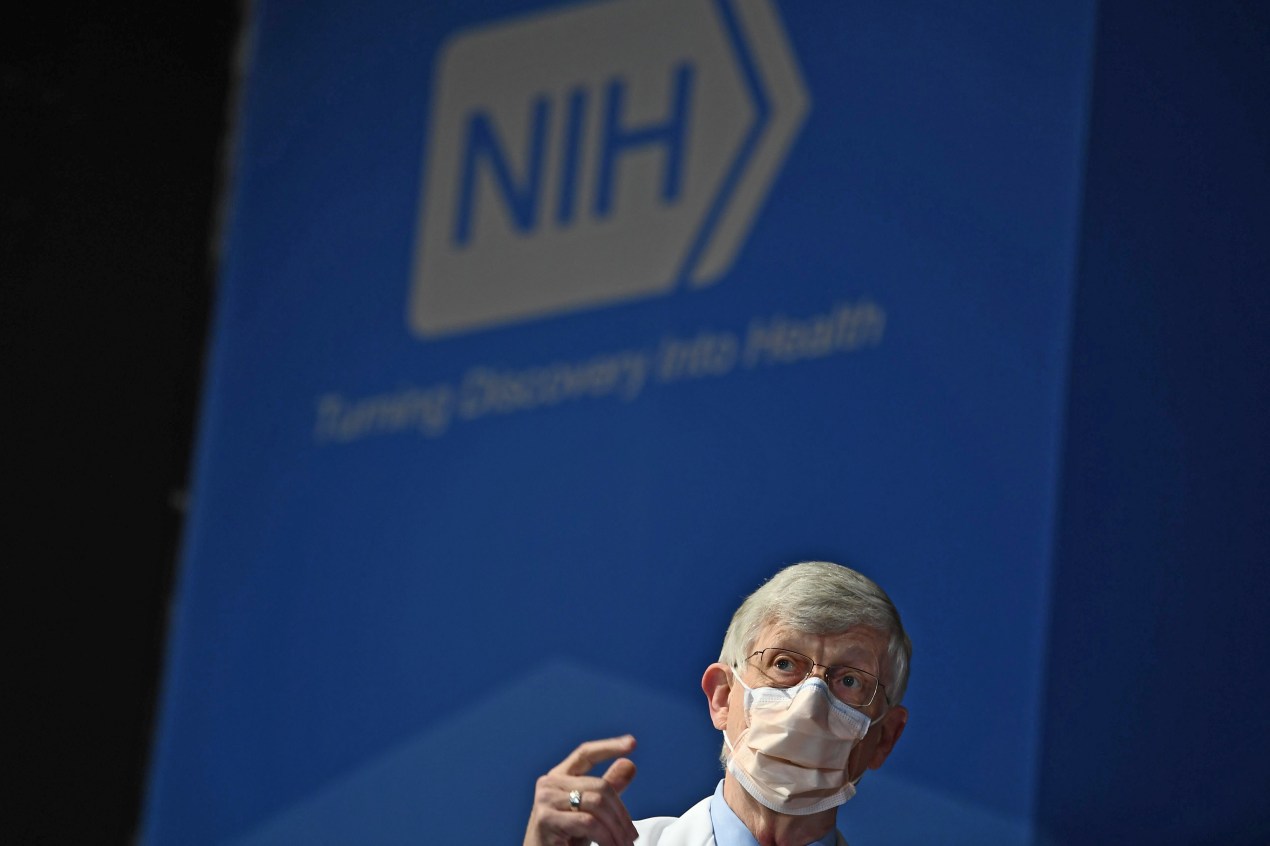 A photo of the former NIH director speaking in front of the NIH logo.
