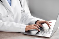 A photo of a doctor in a white coat typing on a laptop.
