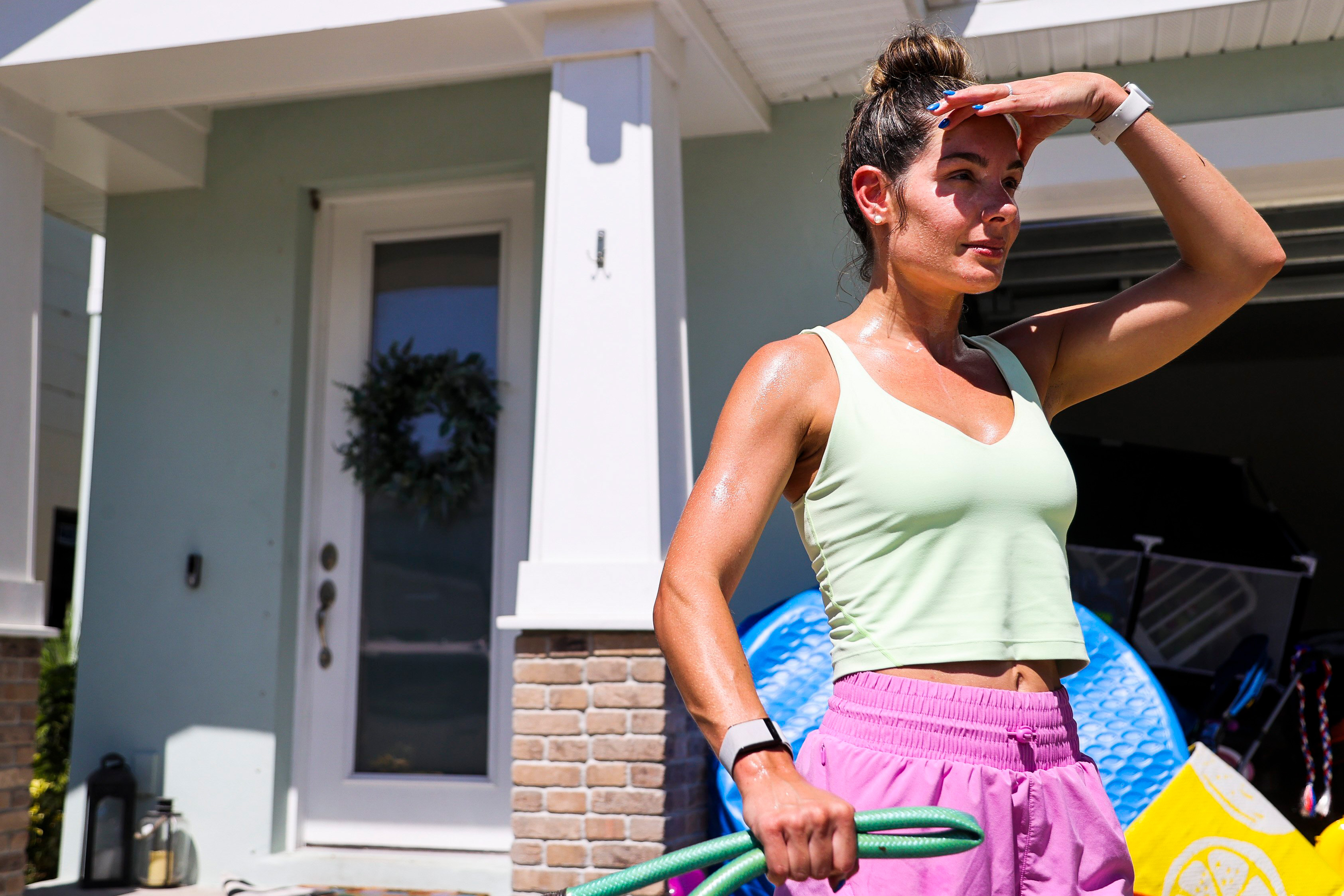 A photo of a woman holding a garden hose outside of her home. She's holding her hand up to shield her eyes from the sunlight.