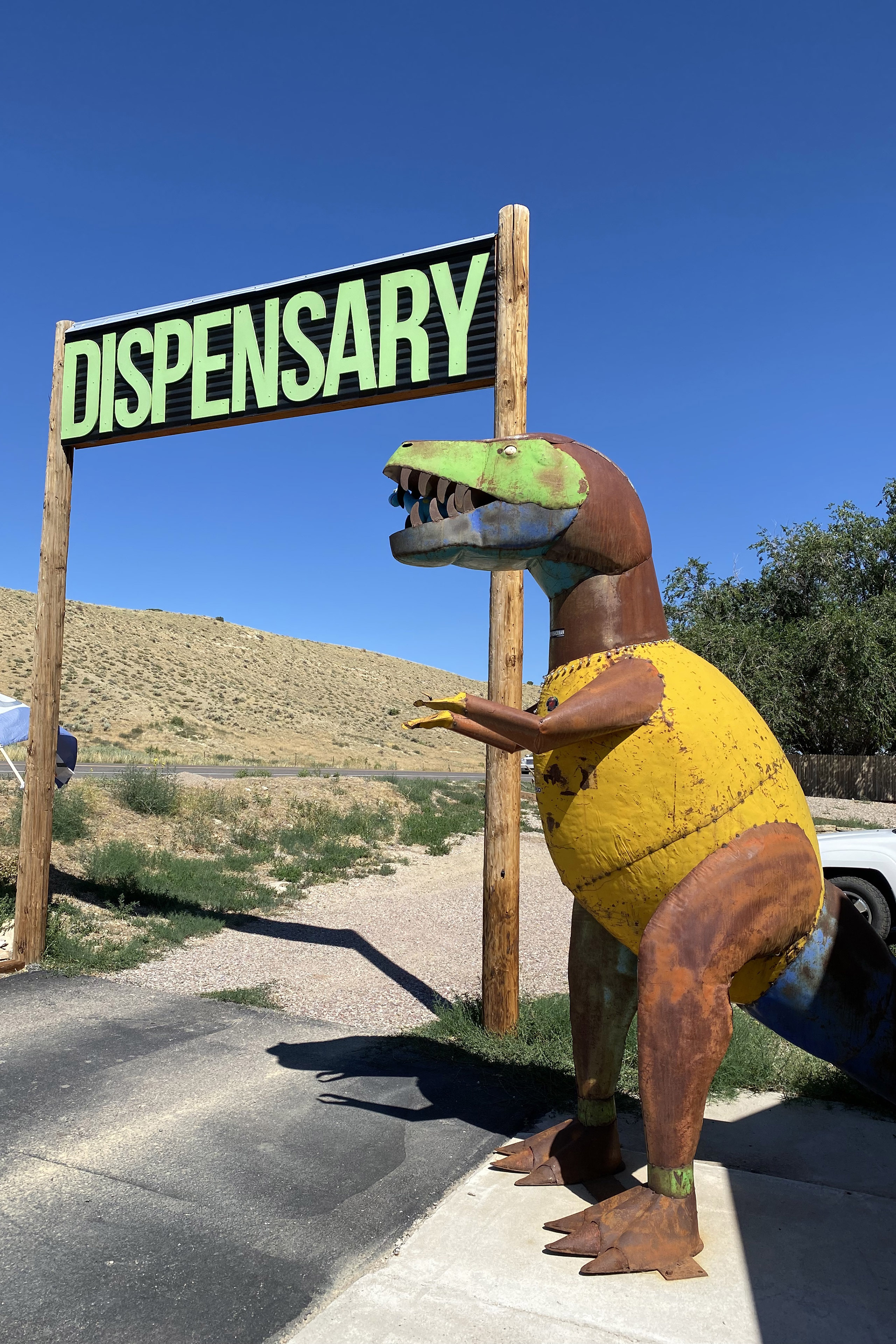 A colorfully painted metal dinosaur, which looks to be approximately over 7 feet tall, stands beside a sign that reads: "DISPENSARY"