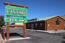 A outdoor large sign reads: "ROCKY MOUNTAIN CANNABIS – Established 2009 – 21+" and points towards a cabin that has a large marijuana leaf on the side.
