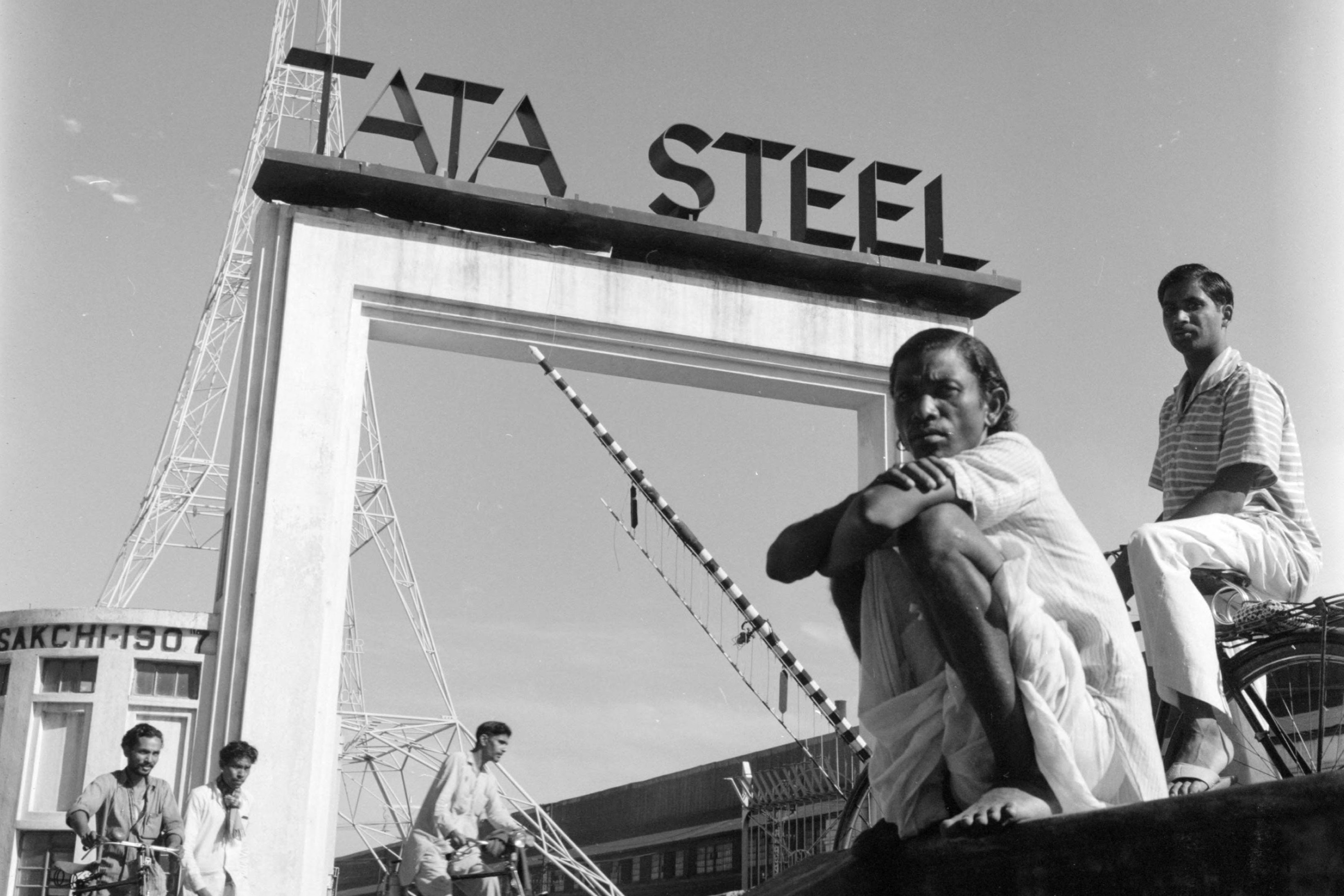 A photograph shows the entrance to Tata Steel in Jamshedpur. There is a large sign that reads, “TATA STEEL.” Two people are sitting below it and looking toward the camera. More people are walking through the entryway in the distance.