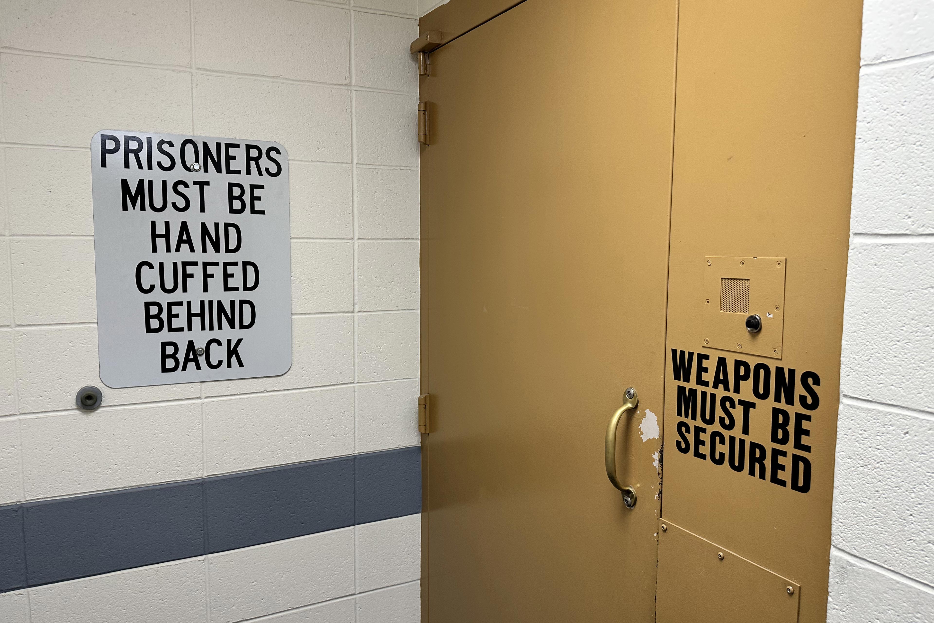 A photo of a sign on a prison wall that reads, "Prisoners must be handcuffed behind back." A door next to it has a sign that reads, "Weapons must be secured."