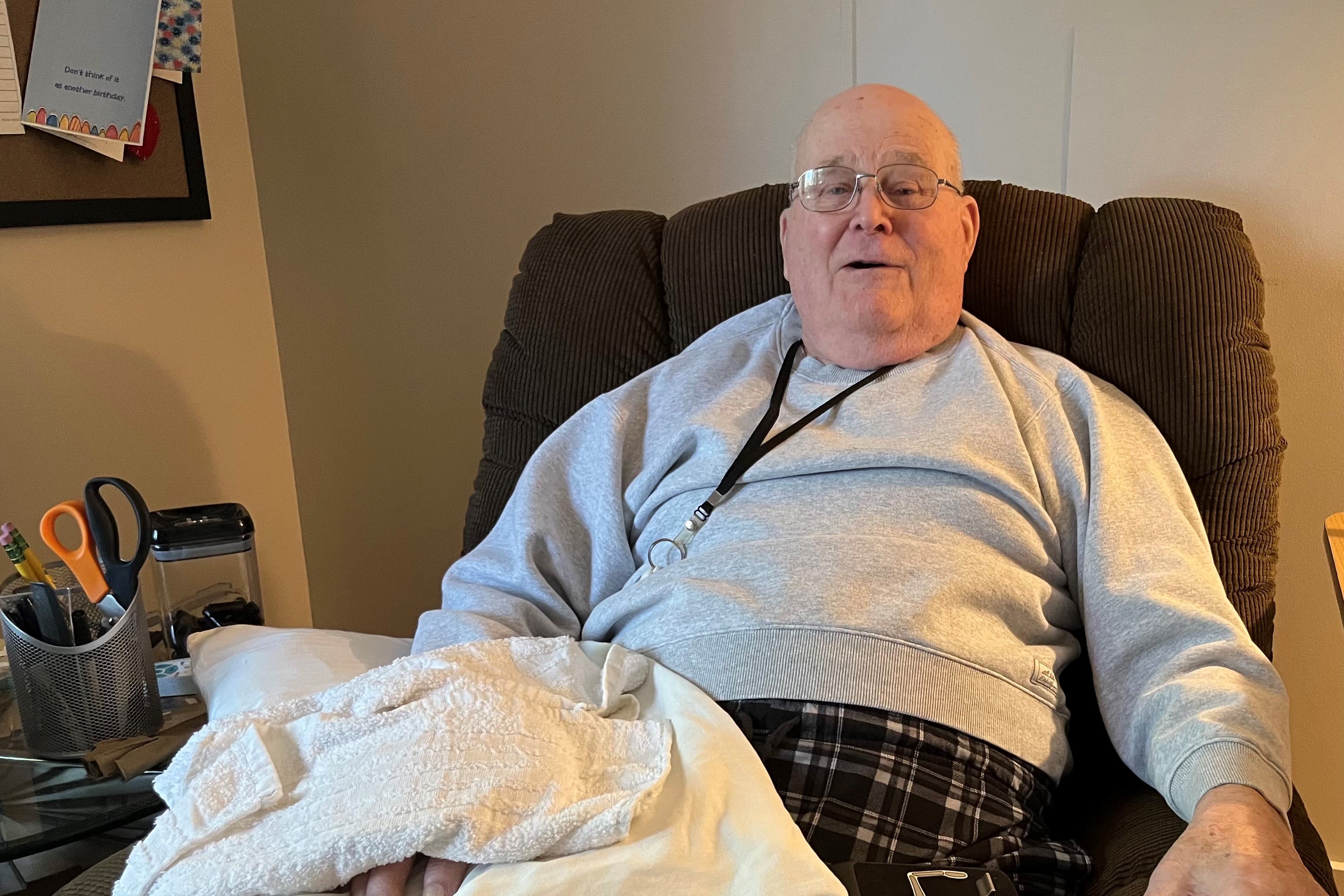 A photo of an older man sitting in a chair inside a nursing home.