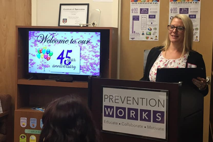 Melanie Witkowski is standing at a podium in a small room. Beside her, a screen reads: "Welcome to our 45th year anniversary". In front of her, a different sign reads: "PREVENTION WORKS – Educate - Collaborate - Motivate"