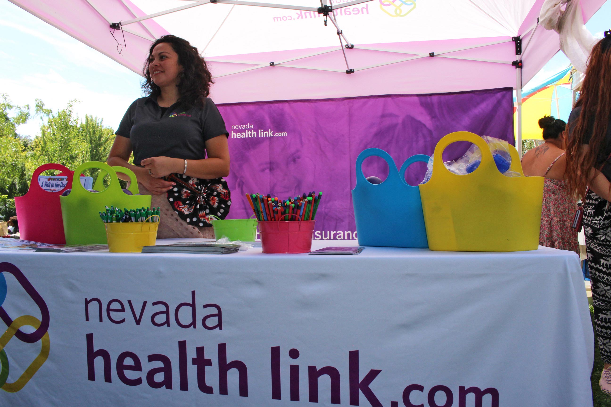 A photo of a woman standing at a booth with with a tablecloth that reads, "nevadahealthlink.com."