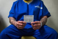 A man in blue prison uniform sits and holds out a card in front of him with both hands.