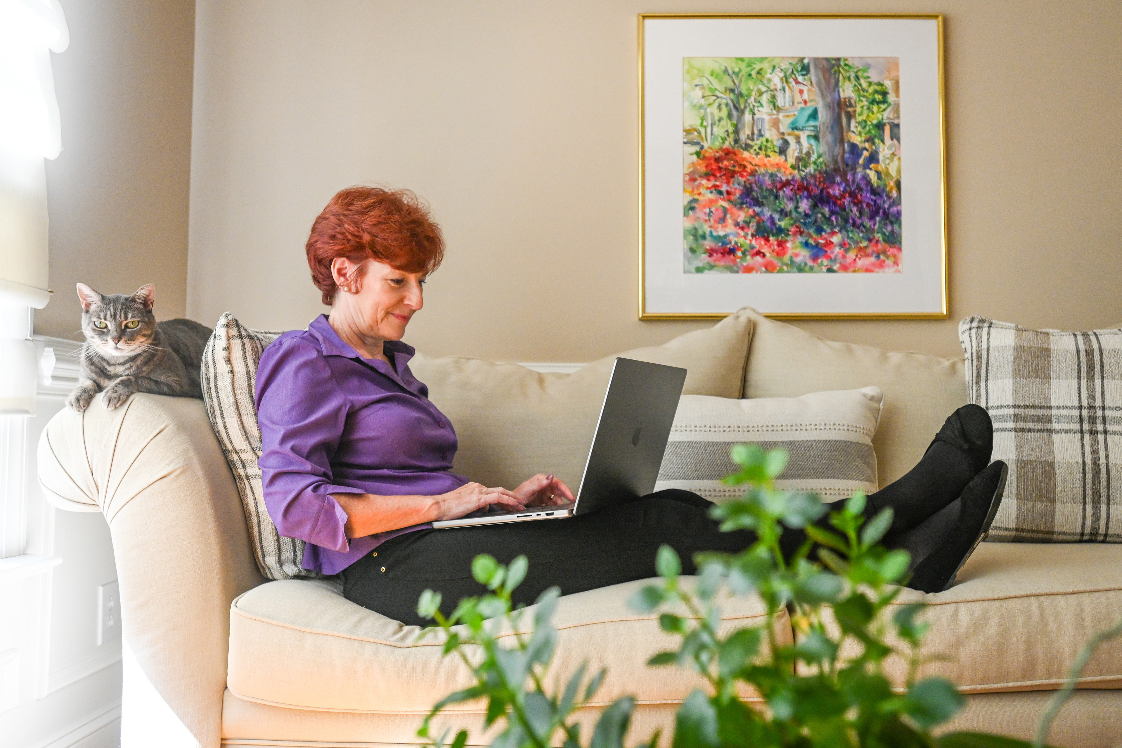 A photo of a woman using a laptop while lounging on a couch next to a cat.