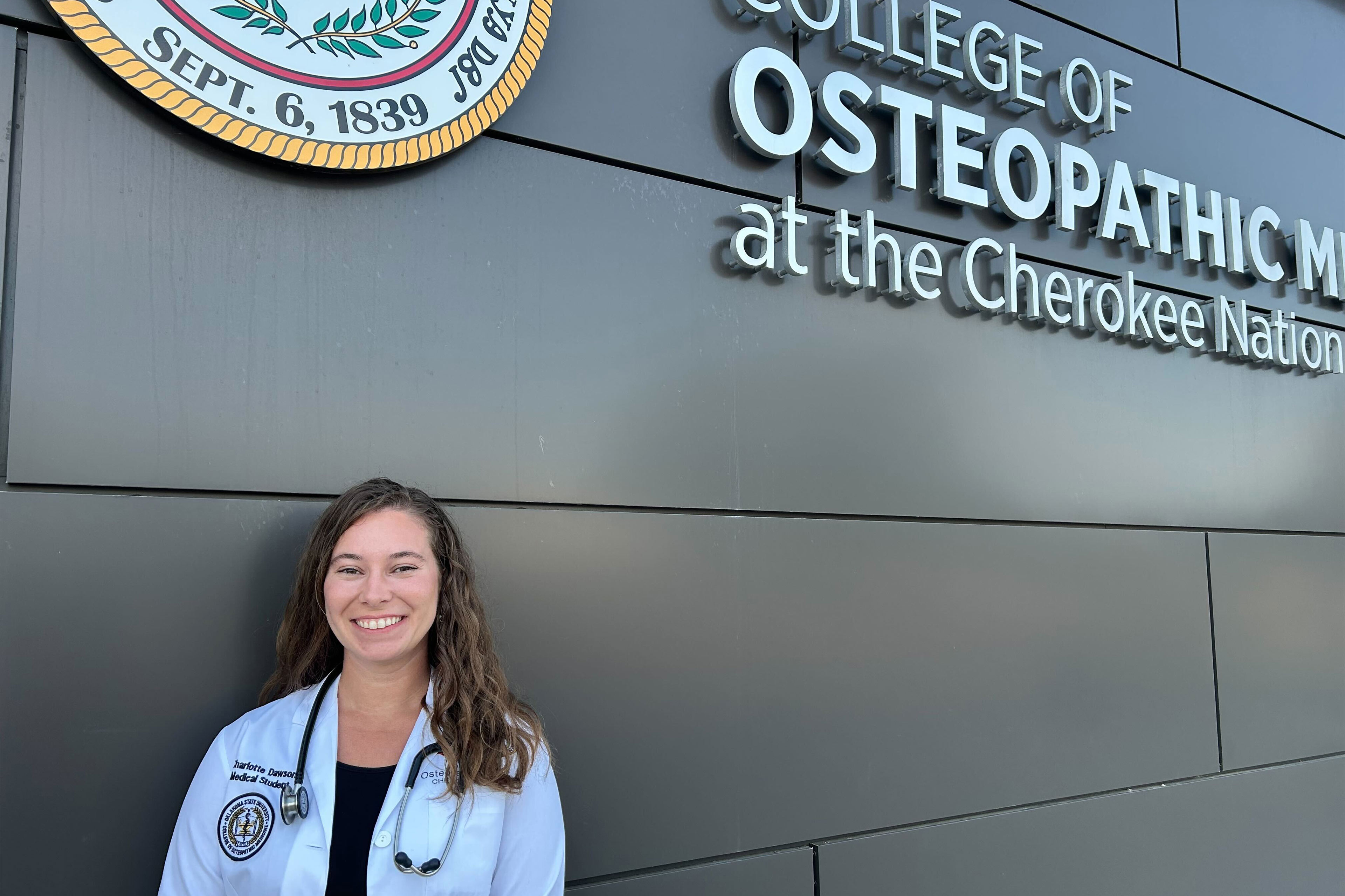 A photo of a medical student smiling. She is standing in front of the medical school's building in Tahlequah, Oklahoma.