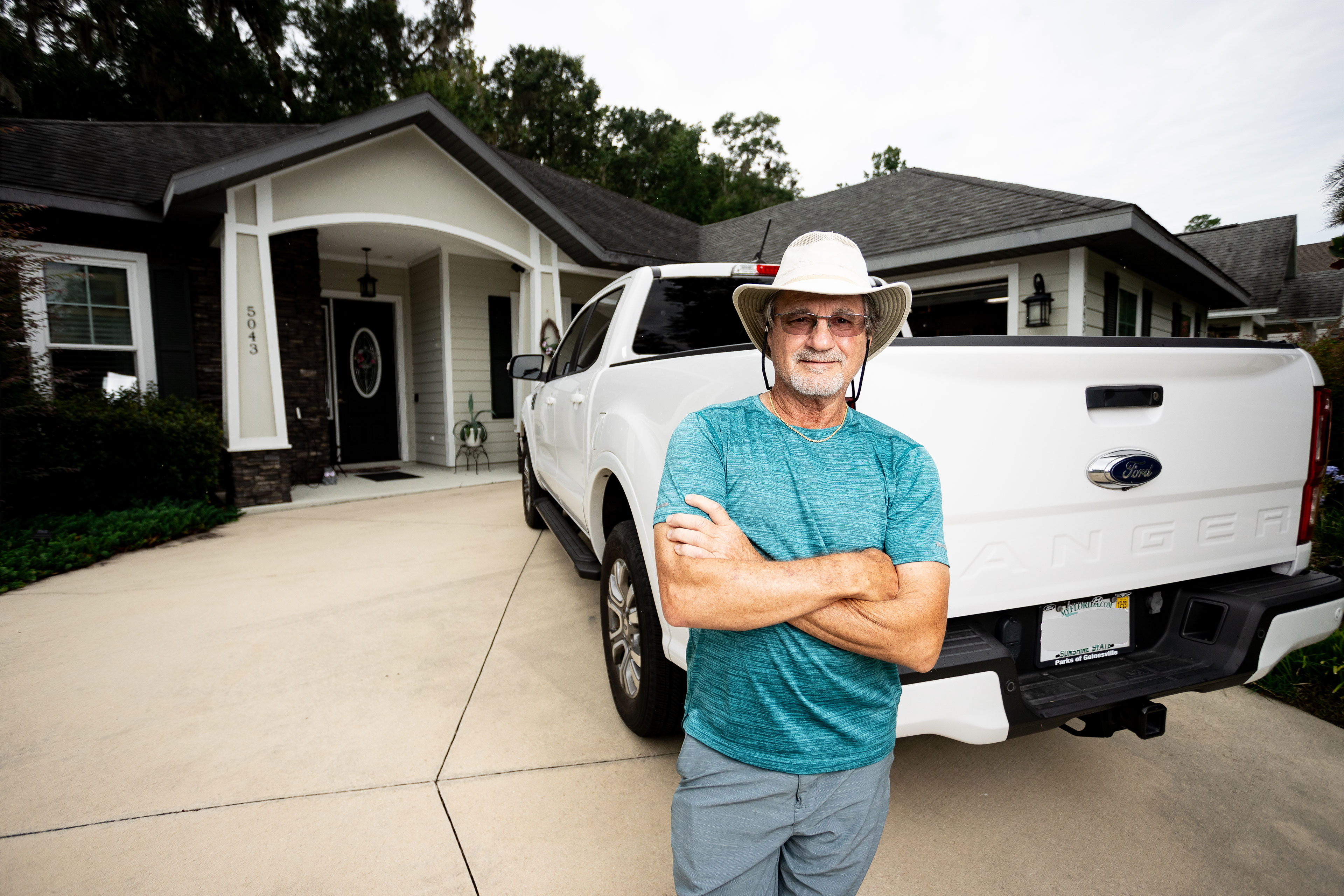 A man with his arms crossed in leans against a white pickup truck parked in a driveway.