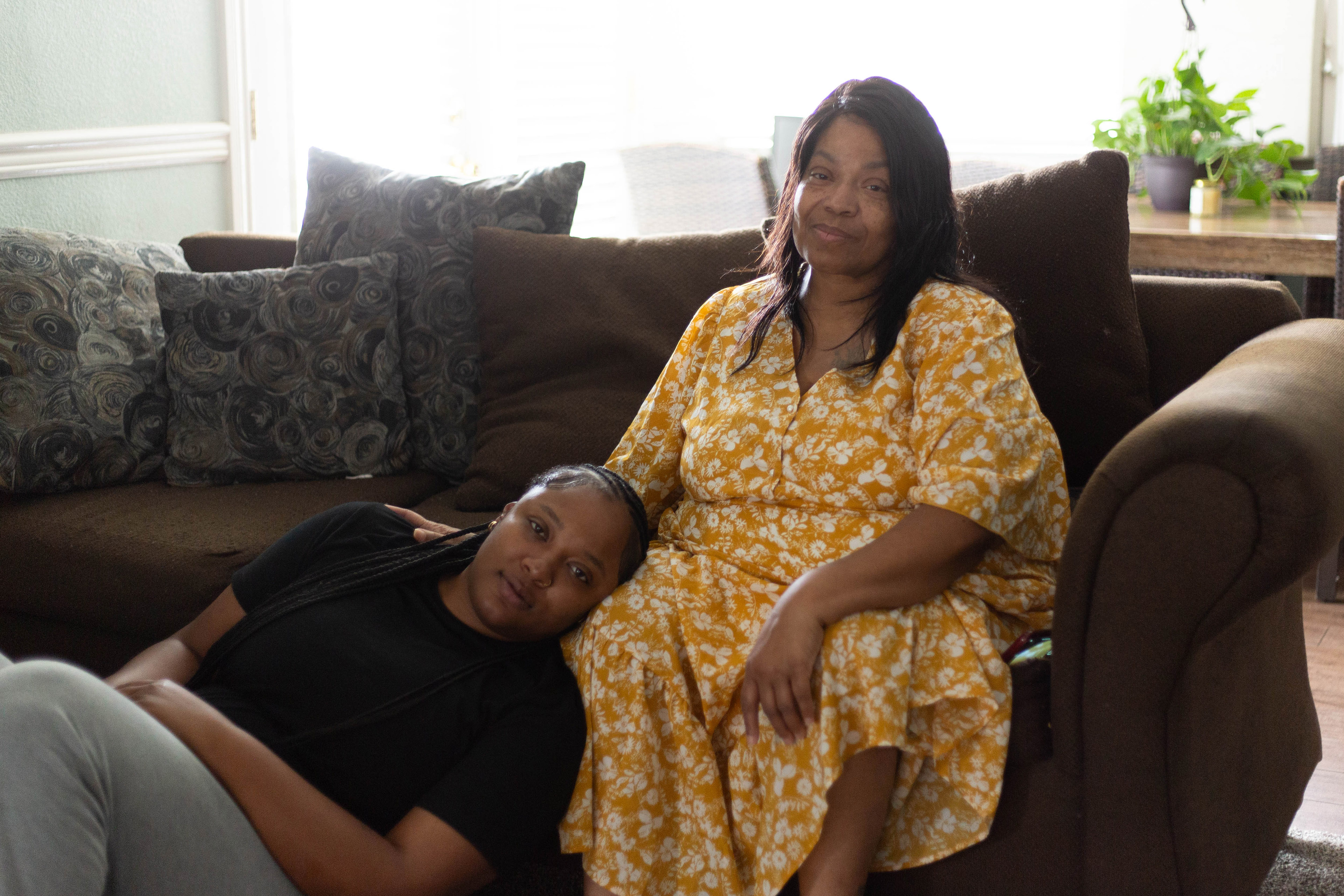 Teresa Johnson is sitting on a couch in her home, with 22-year-old granddaughter Iasia Bailey sitting on the floor beside her. She rests her head on her grandmother's knee.