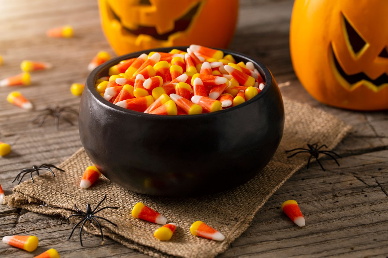 A photo of a bowl of candy corn surrounded by carved pumpkins and small plastic spider decorations.