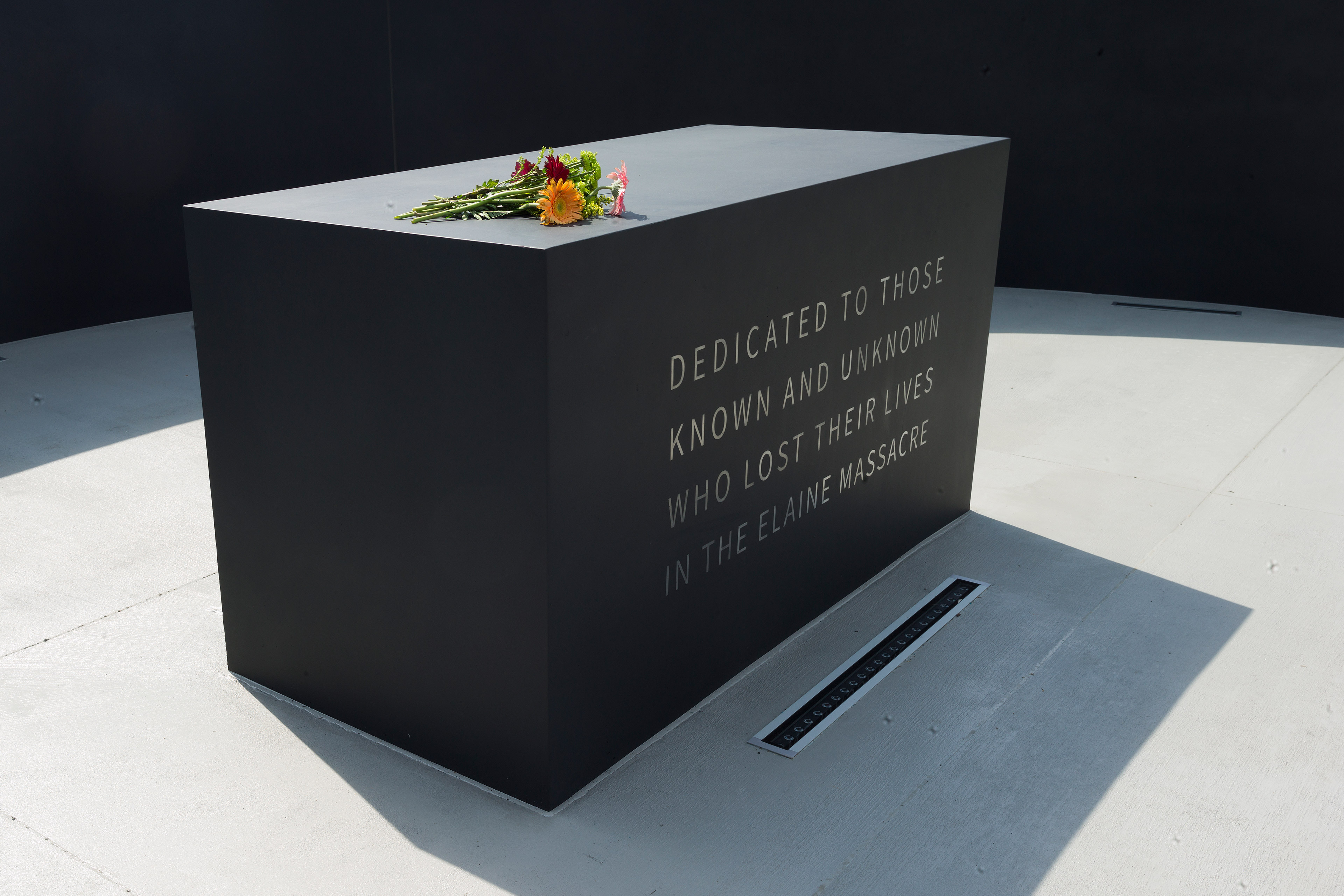 A photo of a memorial with flowers resting on top of it. An inscription on it reads, "Dedicated to those known and unknown who lost their lives in the Elaine massacre."