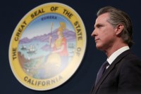 A photo of Governor Gavin Newsom during a press conference taken from the side. On the wall behind him is California's state seal.