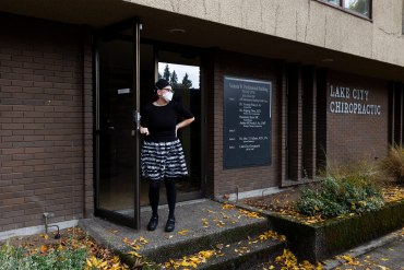 A photo of a medical assistant wearing a face mask holding a door open while she waits outside.