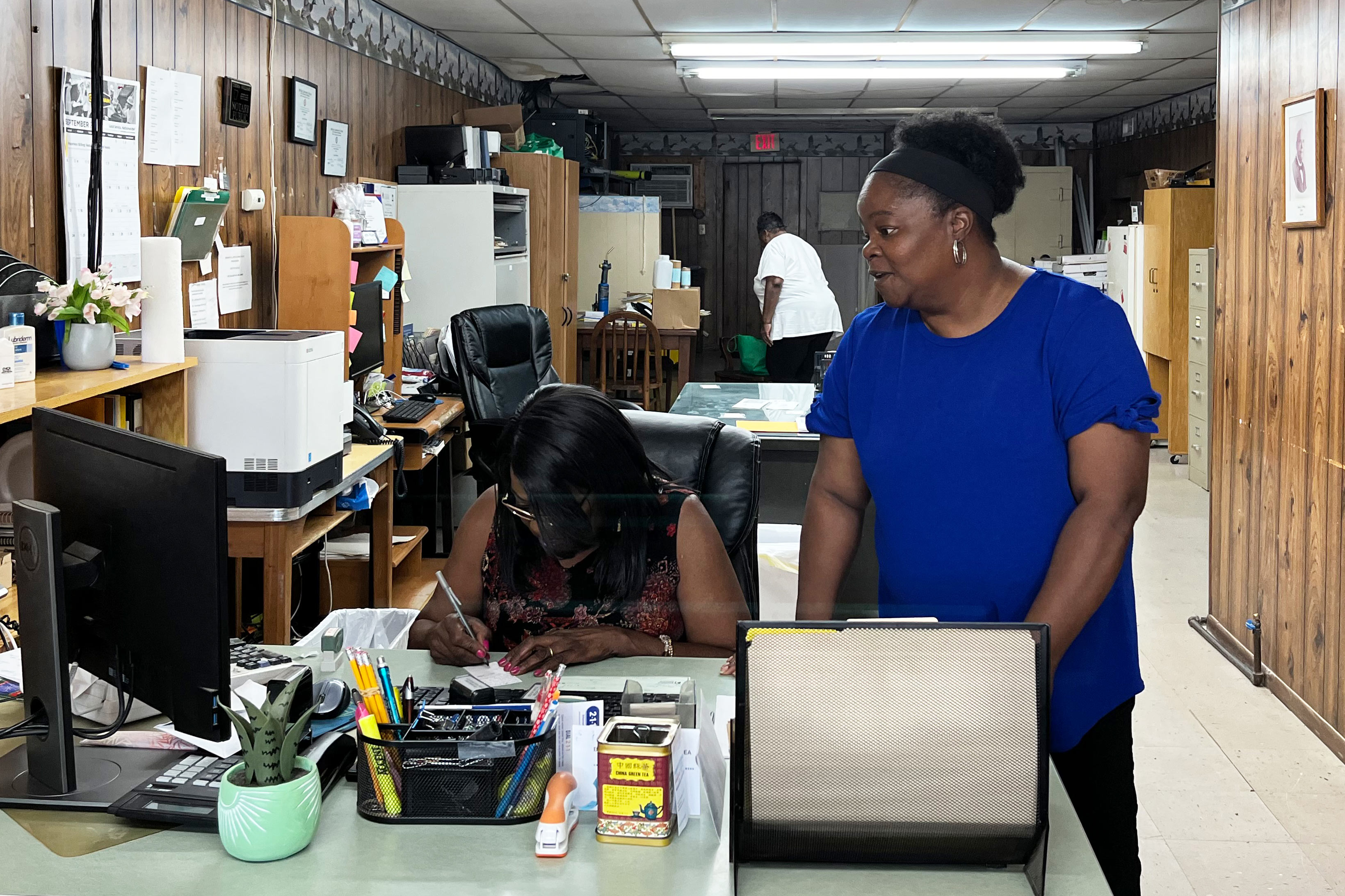 A photo of a woman standing indoors and looking to another woman to her right working at a computer.