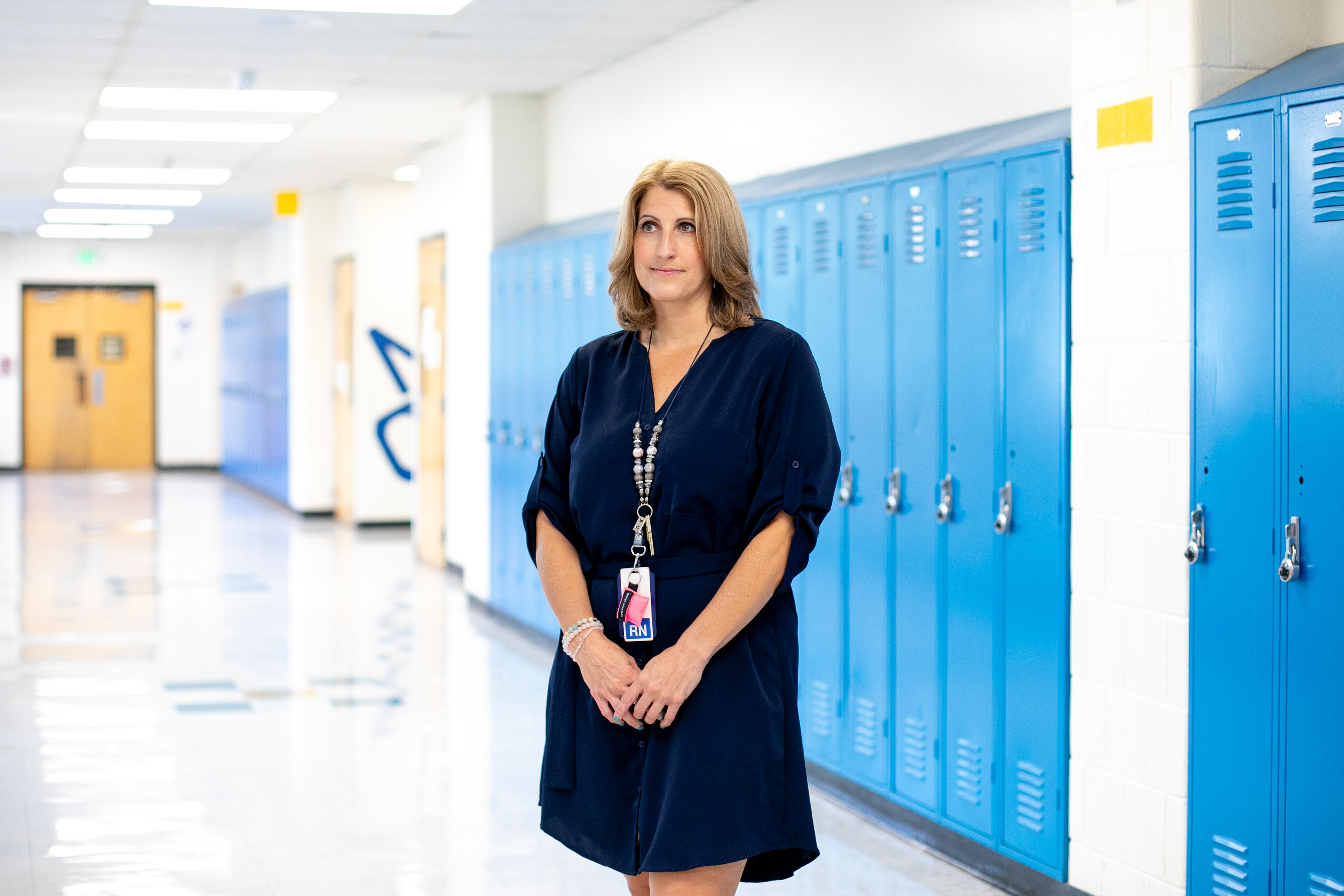 A woman in a dark dress stands in a school hallway with blue lockers behind her.