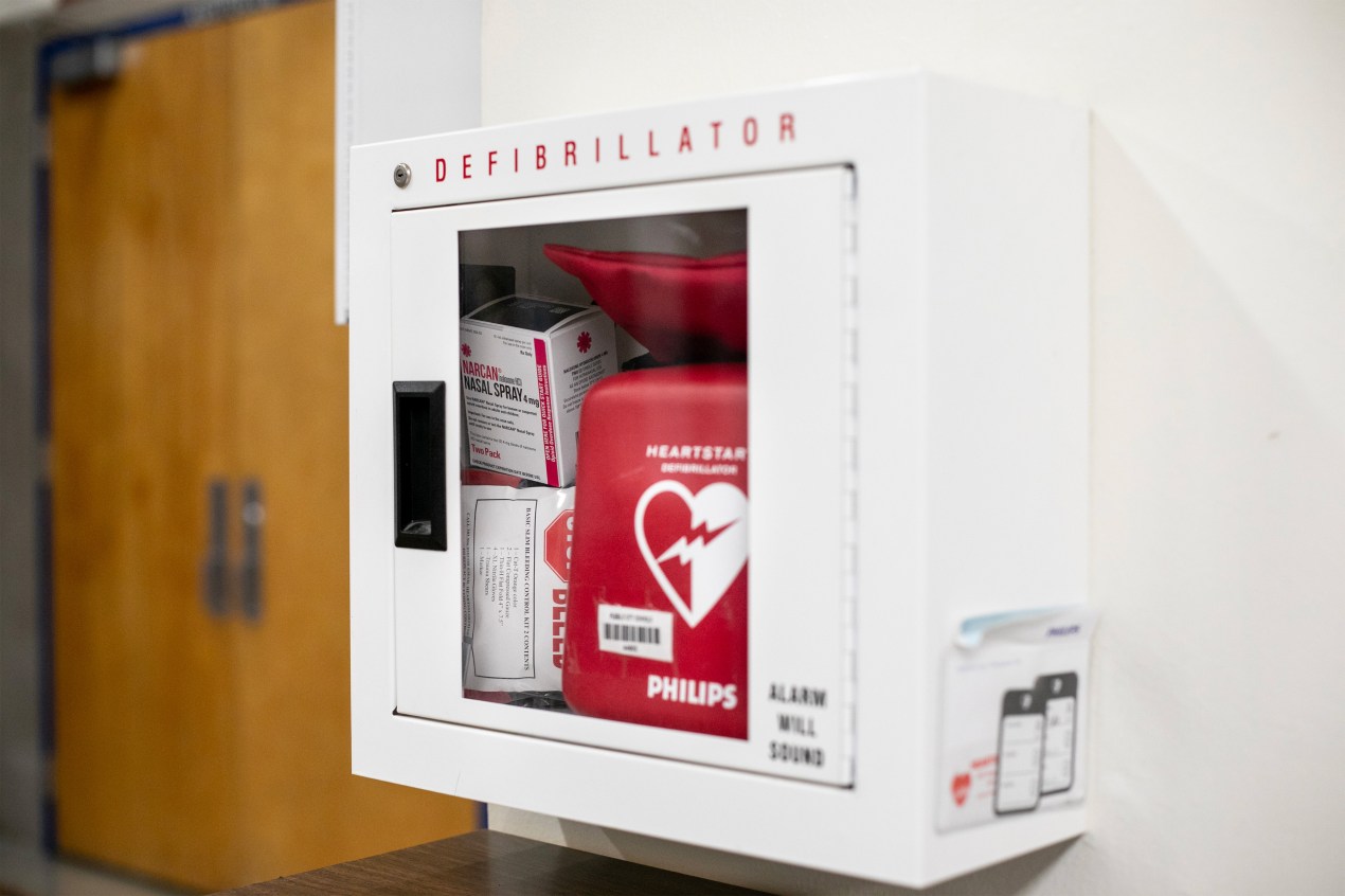 An image of a cabinet on a wall with a glass door and a box reading Narcan and a defibrillator inside.