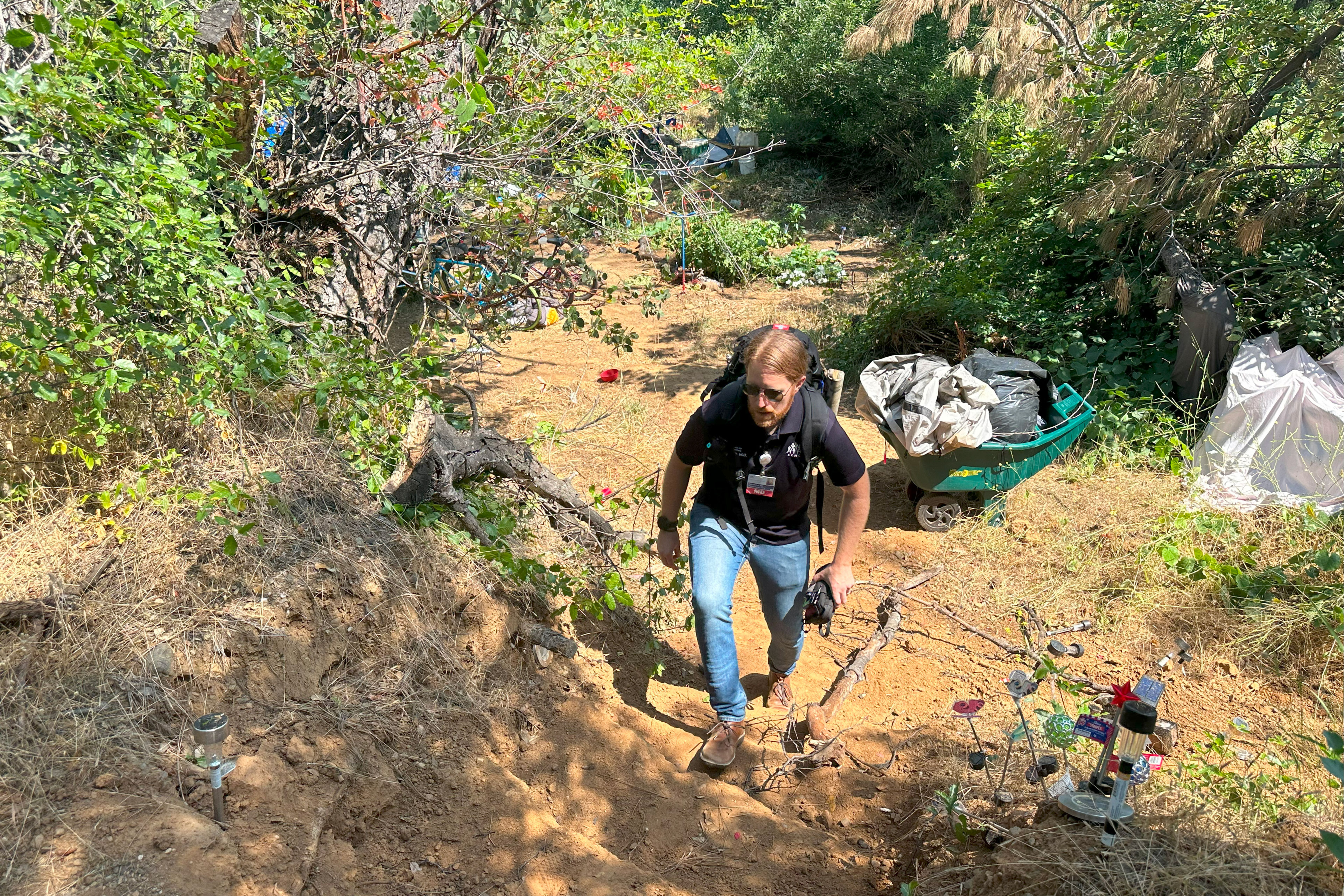 A man wearing a backpack begins to climb a flight of dirt stairs out of an encampment.