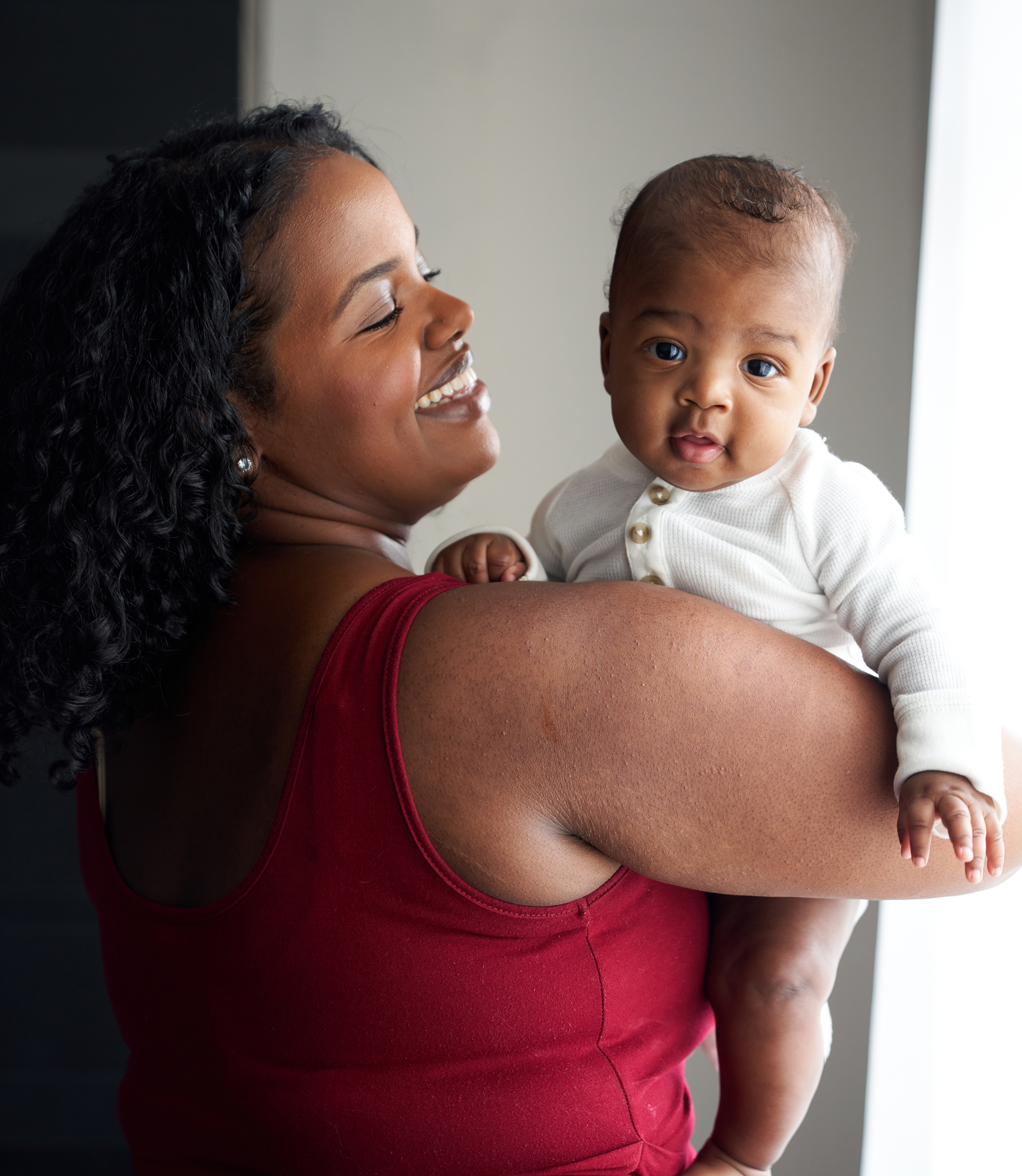 A woman in a red dress stands while holding her baby, smiling at him as he smiles at the camera