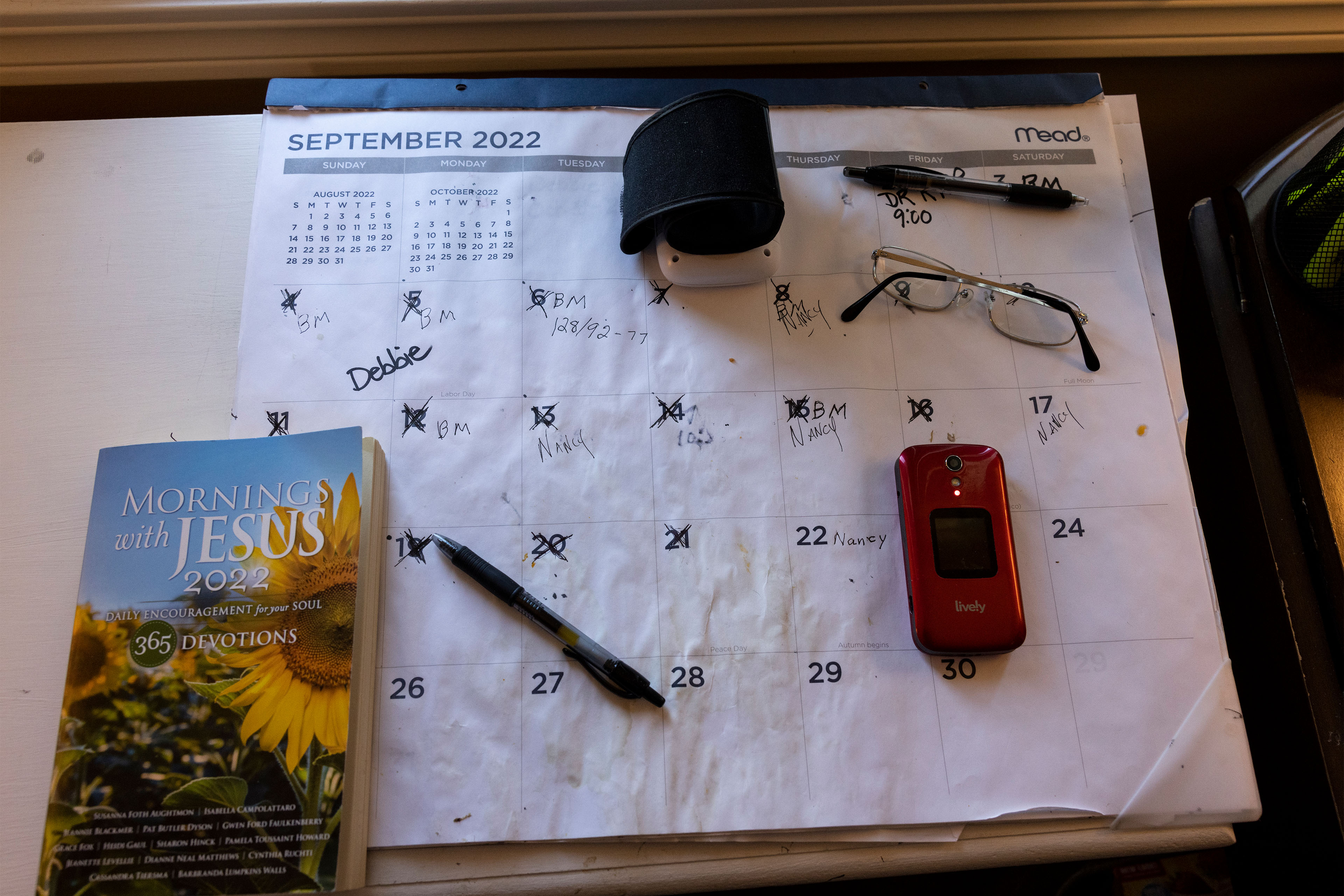 A photo of a calendar, a pair of glasses, a devotional book, a cell phone, and some pens resting on a table.