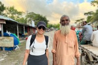 Céline Gounder, wearing a baseball cap, sunglasses, white T-shirt, and backpack, stands beside Delowar Hossain, a former smallpox eradication worker. He has a long white beard and wears a loose peach-colored long-sleeved shirt. They both smile at the camera. In the background, a sunset highlights pillowy clouds in warm shades of pale yellow that contrast with bits of blue sky.