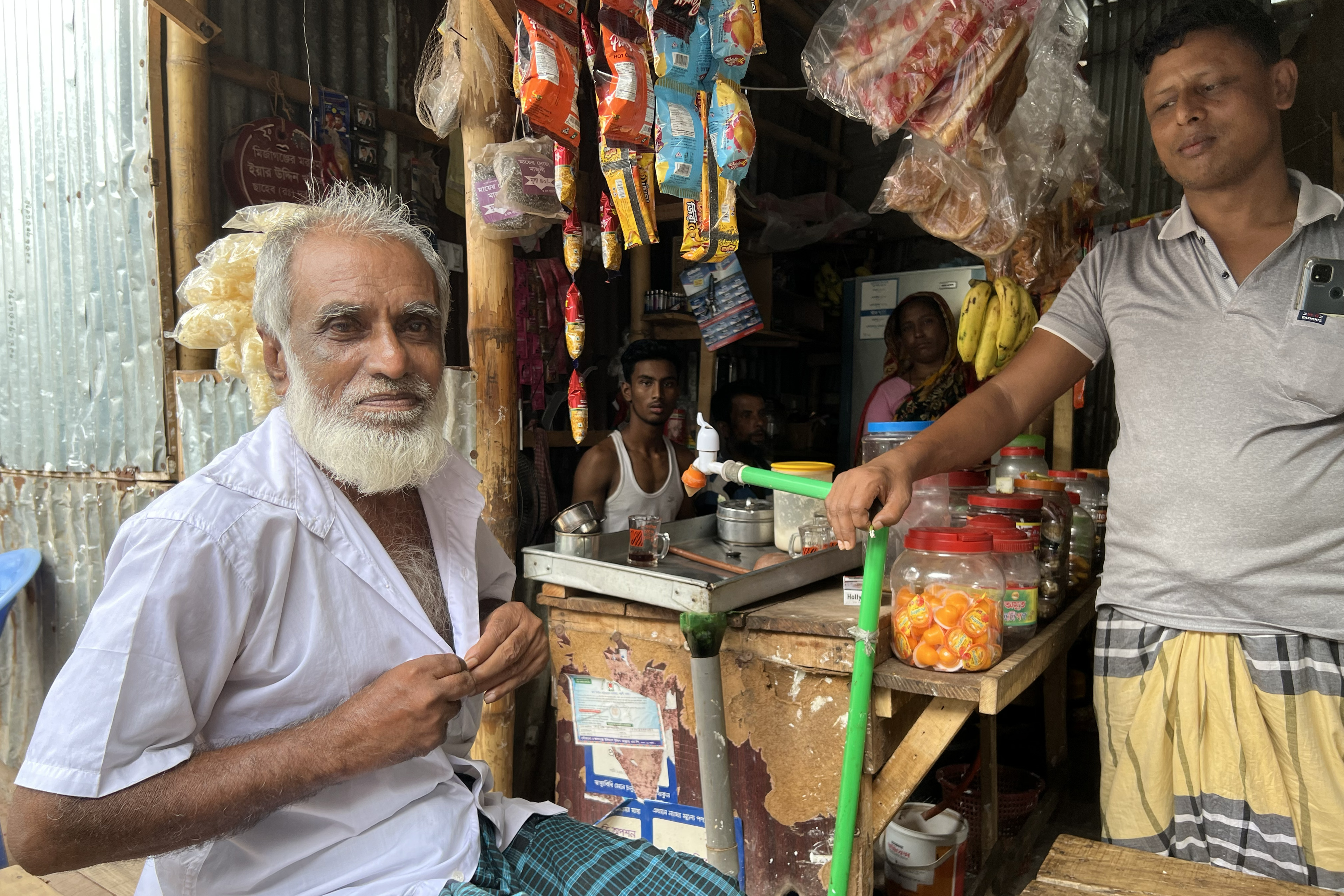Shohrab, a man in his early 70s, sits outside a tea stall on a well-worn wooden bench near his home in Dhaka. Inside the stall, a colorful display of snacks and sweets hang from the ceiling. Three other men and one woman are nearby in the tea stall.