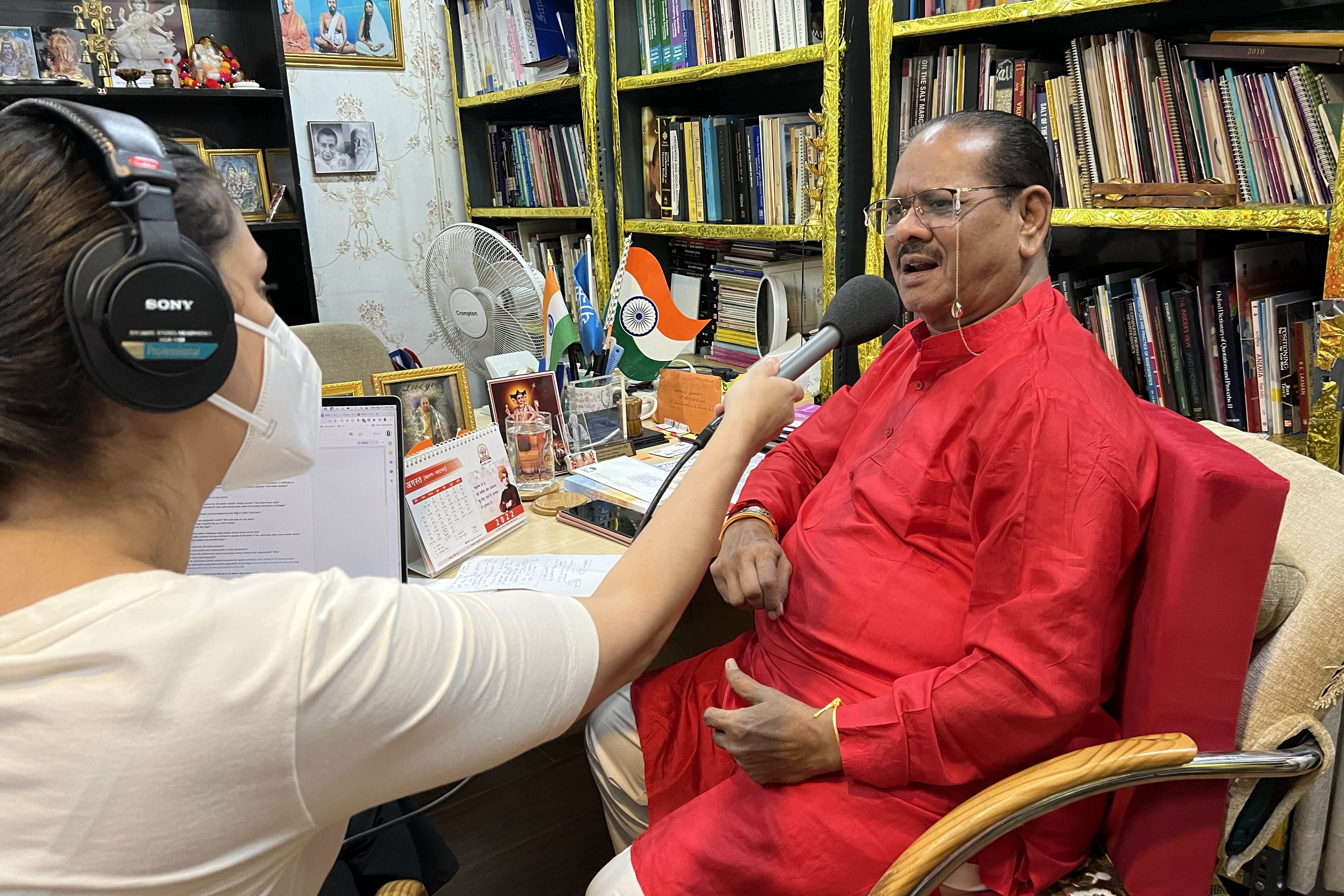 Chandrakant Pandav, wearing a bright red outfit and glasses, off of which hangs a small gold chain that links around the back of his neck, sits in his office as he is interviewed by Céline Gounder. She is to his left, holding a microphone to him as he speaks. She wears professional audio-recording headphones and a KN95 mask.
