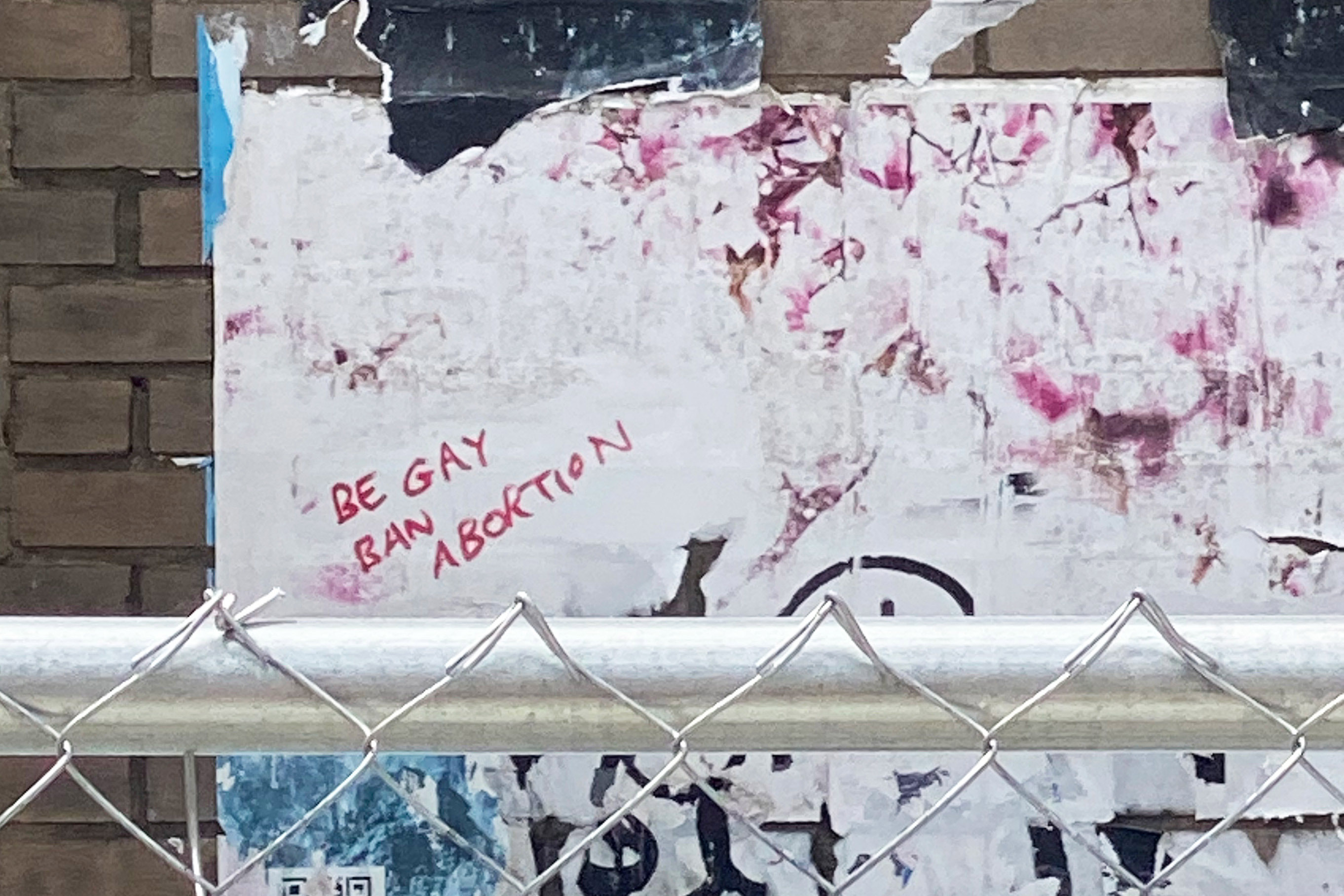 A photograph of graffiti written in red marker behind a chain-link fence. It reads, "be gay / ban abortion."