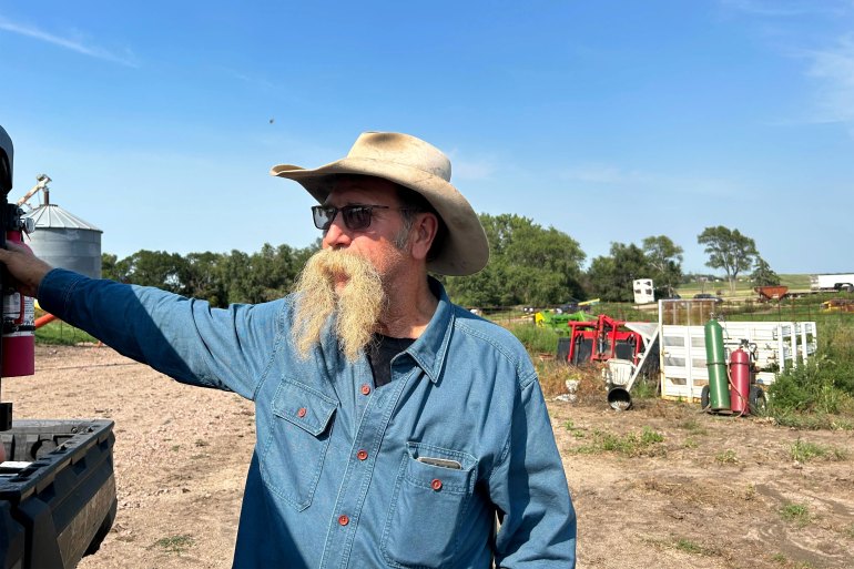 A man with a long mustache and wearing sunglasses and a cowboy hat stands outside and looks off-camera to his right.