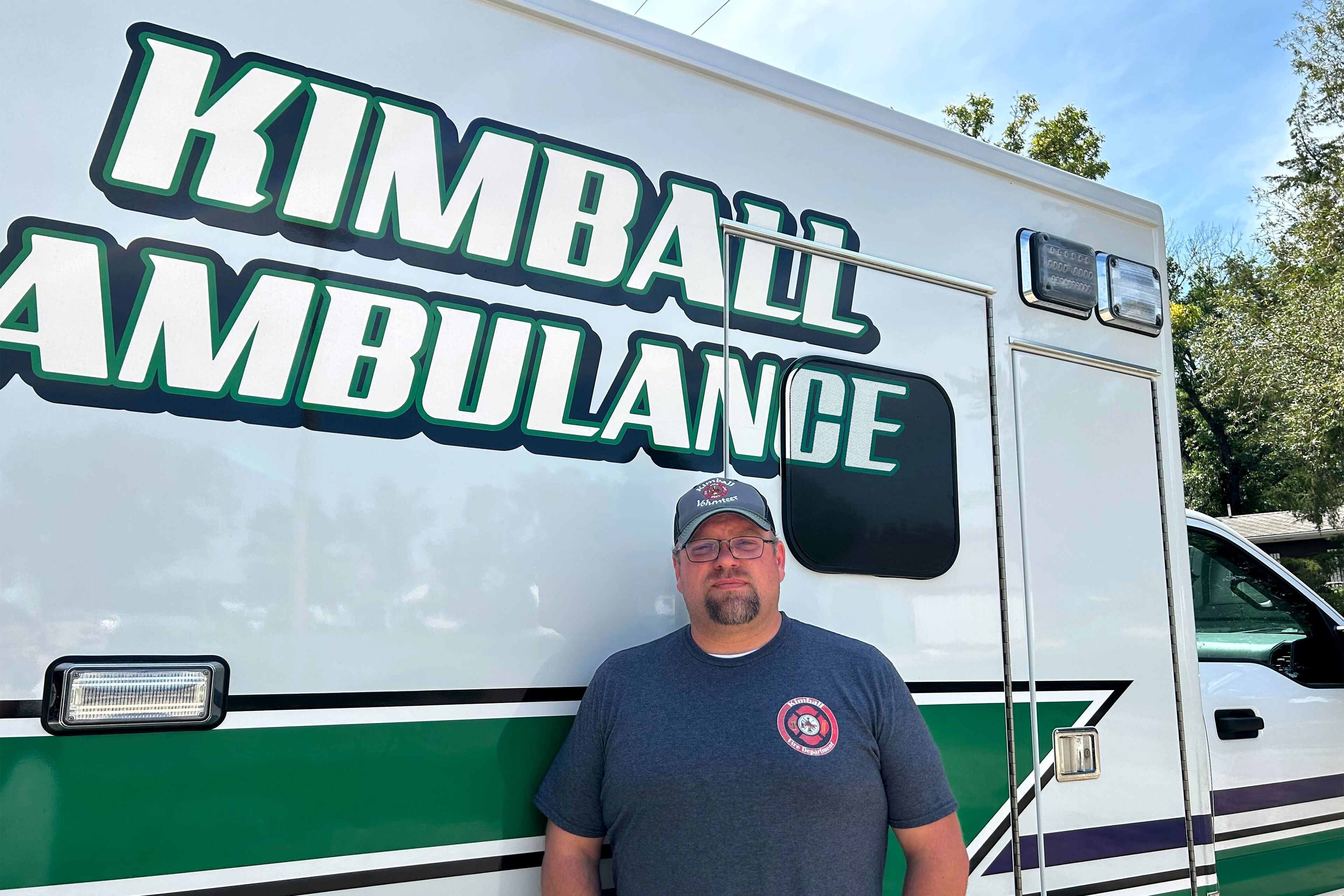 A man wearing a cap stands in front of an ambulance painted with "Kimball Ambulance" on the side.