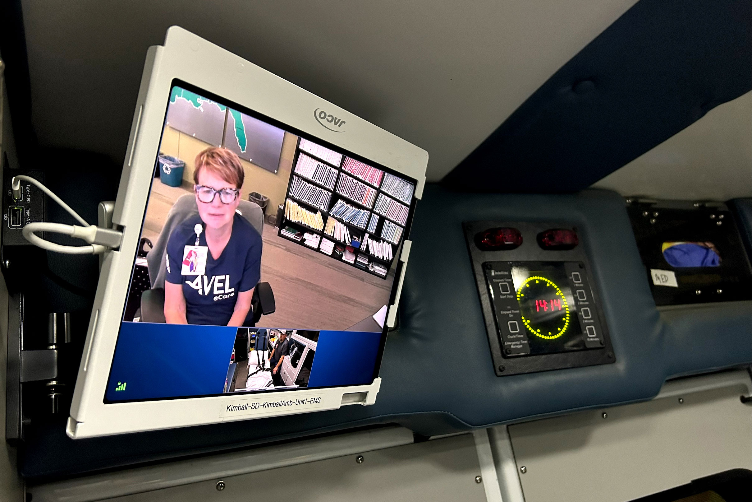 A tablet is mounted on the inside of an ambulance and you can see someone and someone sitting in a chair in an office and wearing a name badge and t-shirt that reads "Avel eCare" is shown on the screen.