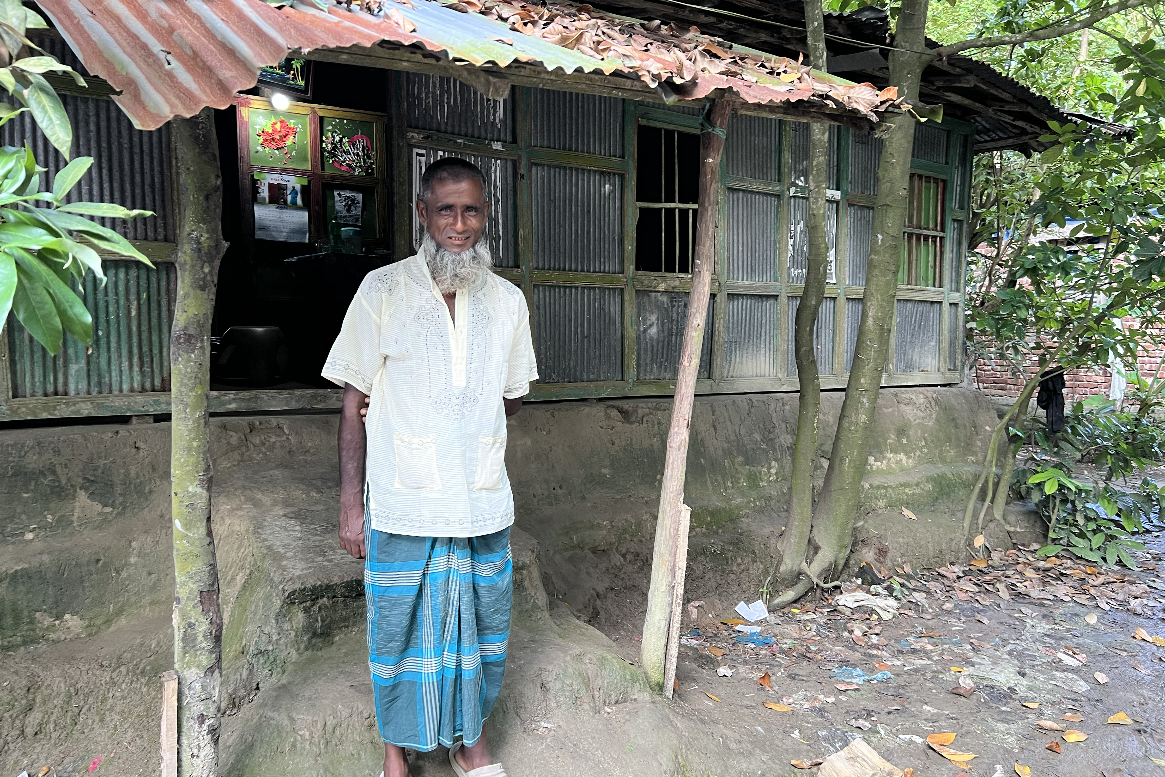 Rahima Banu’s husband stands in front of their home in August 2022. He has a long beard, mostly gray, and wears a daffodil-yellow short-sleeved shirt with a sky-blue garment beneath it. The house is made from bamboo and corrugated metal. The mud stairs that lead inside are dotted with moss.