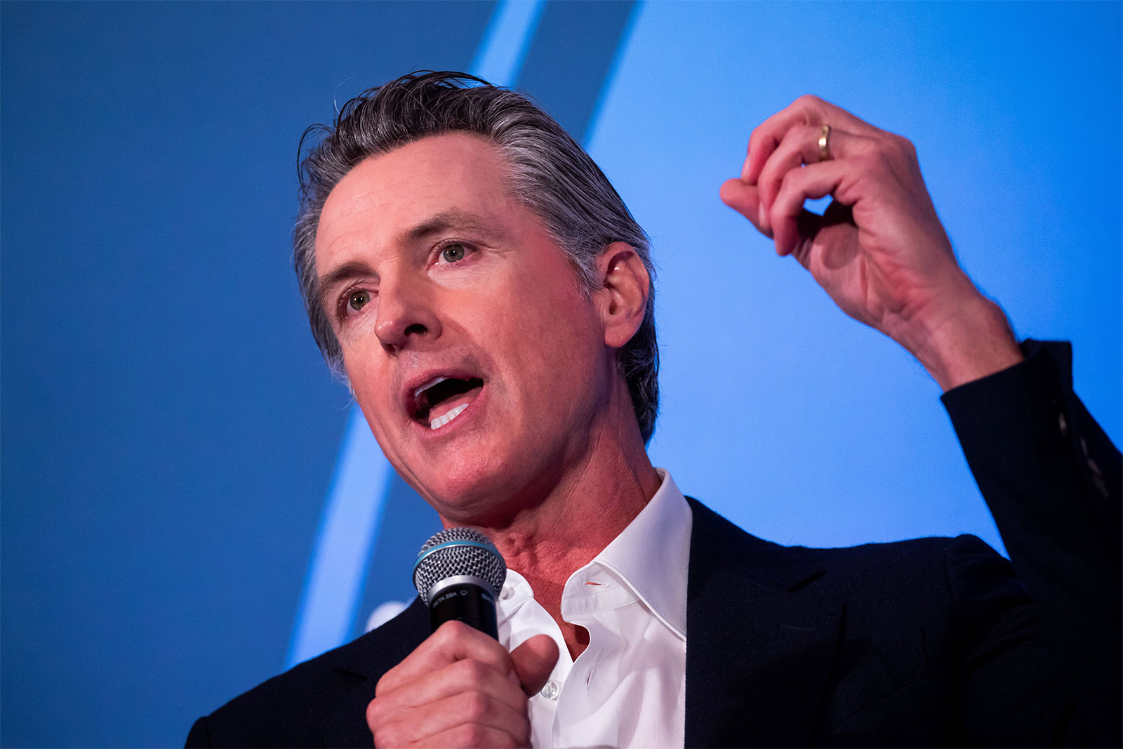 health California Gov. Gavin Newsom speaks into a microphone held in his suitable hand and gestures with his left hand.