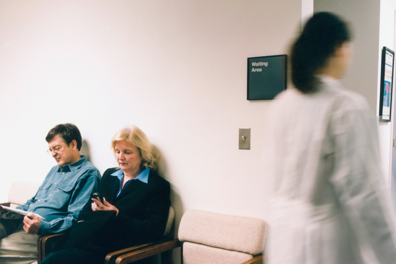Two patients wait in a waiting room at a doctor's office. On the far left, a physician is walking by.