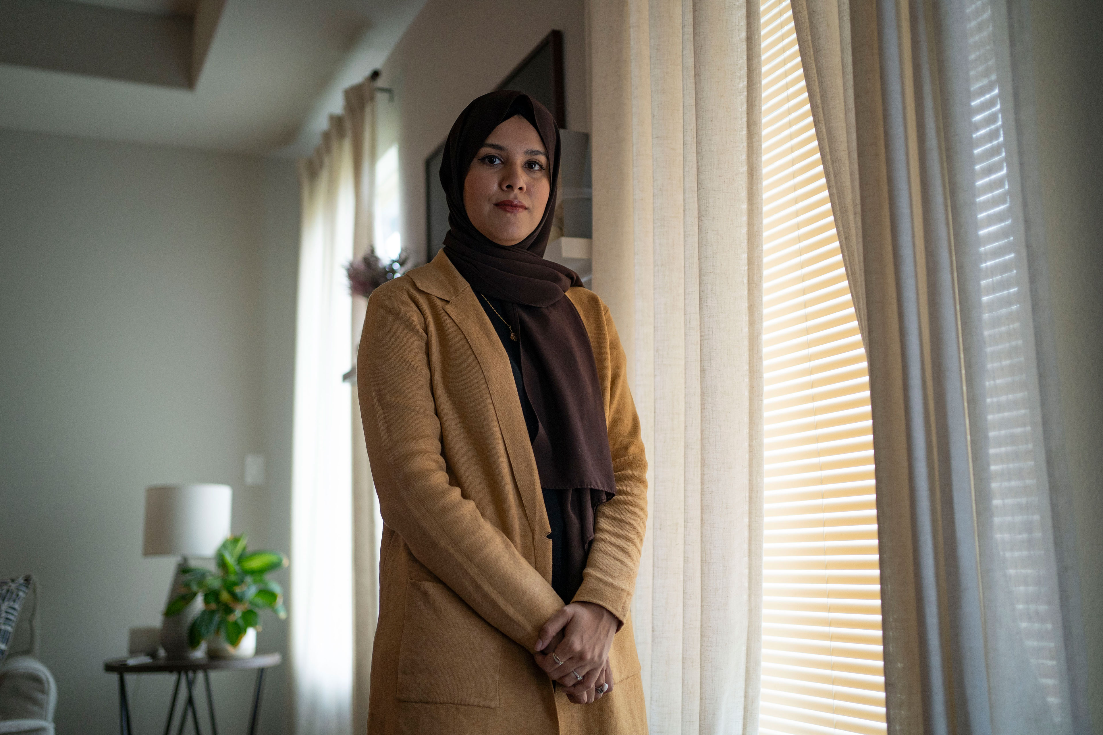 A woman stands by the window looking into the camera. 