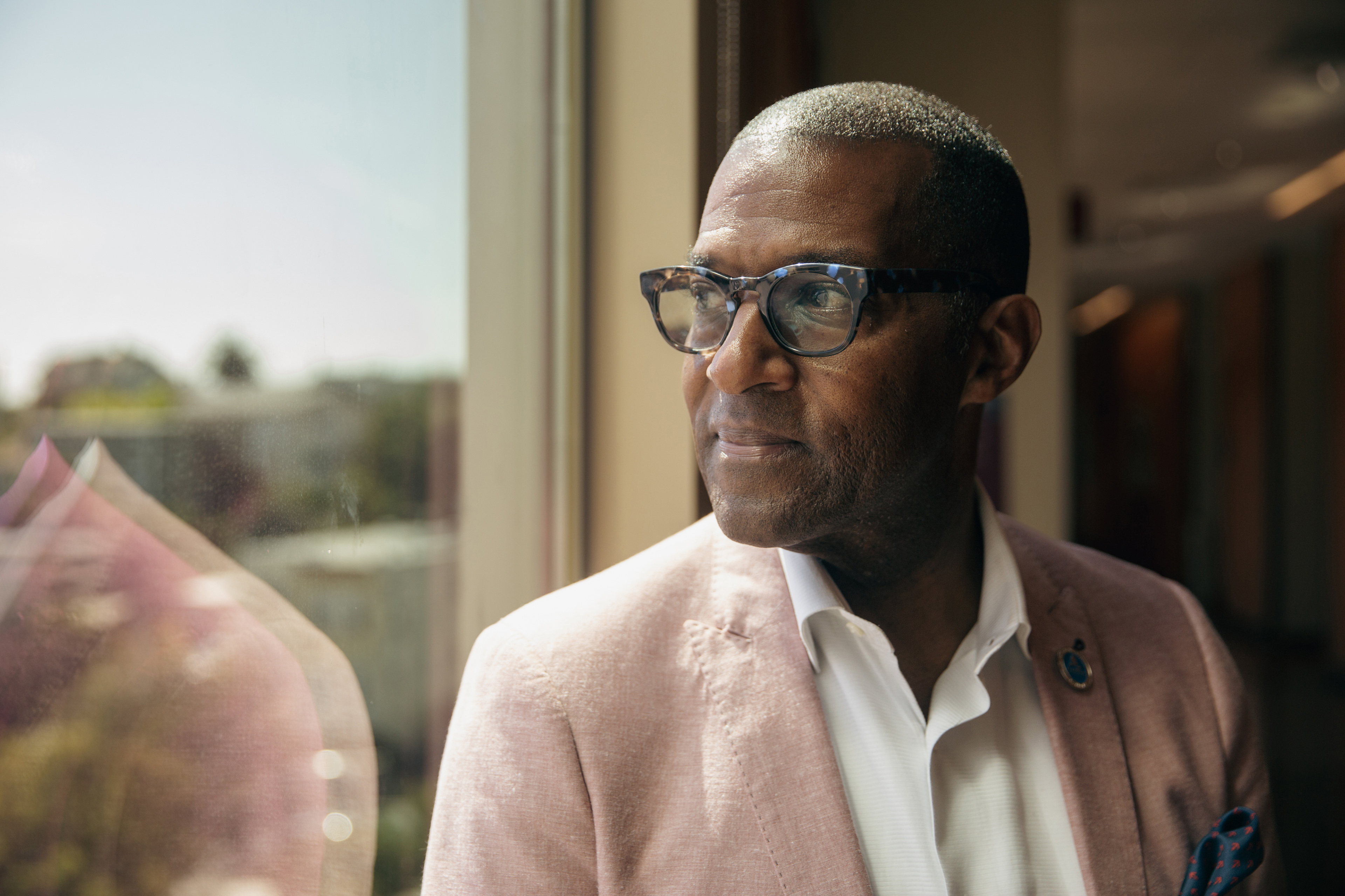 A man stands beside and looks out a large sunny window. He wears glasses and a blazer over his collared shirt.