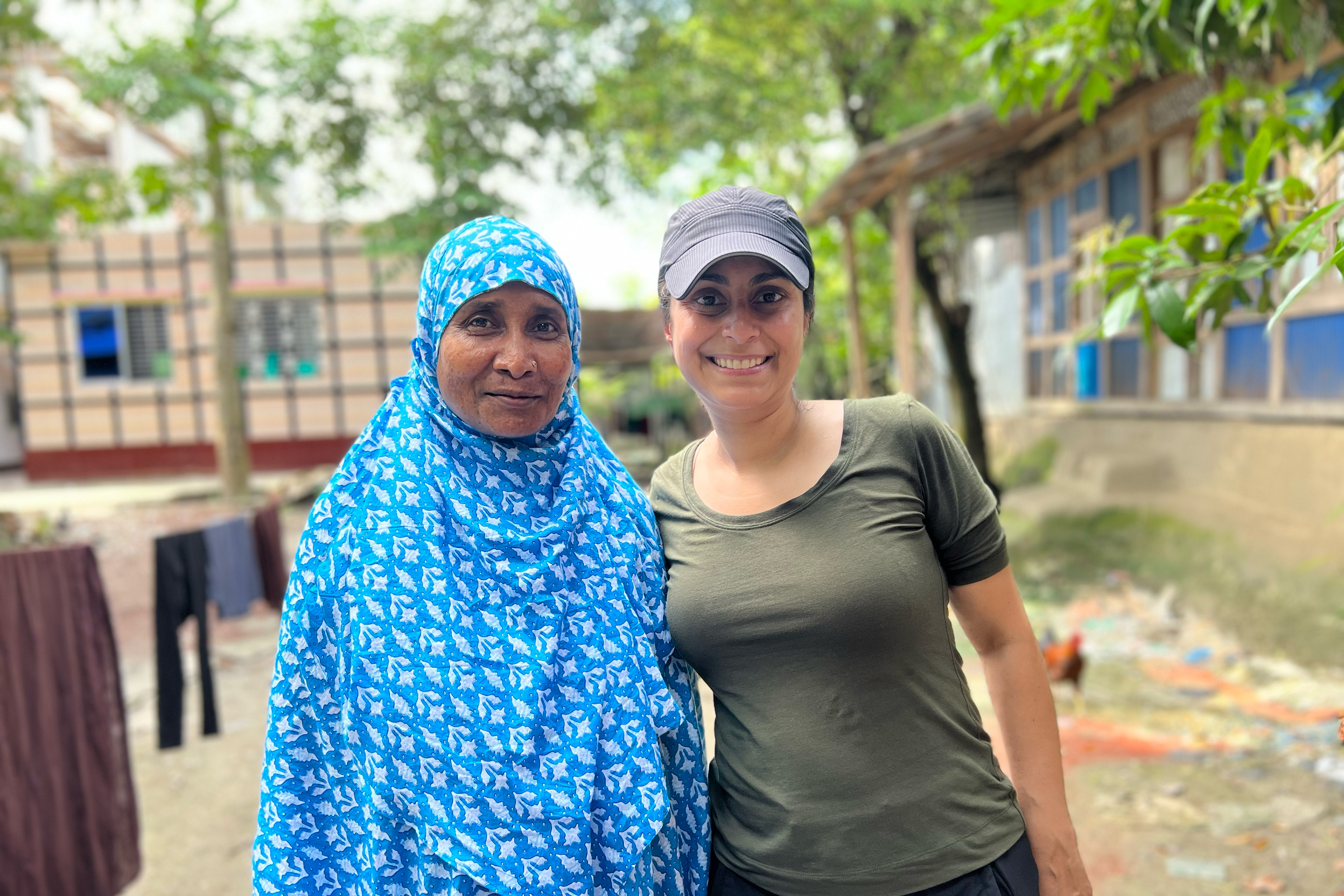 Rahima Banu (left) wears a blue scarf patterned with white flowers. It is draped over her head and body and tucked under her chin. She stands beside Céline Gounder (right), who wears a black baseball cap and green T-shirt. They are in a small courtyard just outside Banu’s home. Laundry dries on a line in the background. Bright green, waxy leaves from growing trees provide shade and softened sunlight.