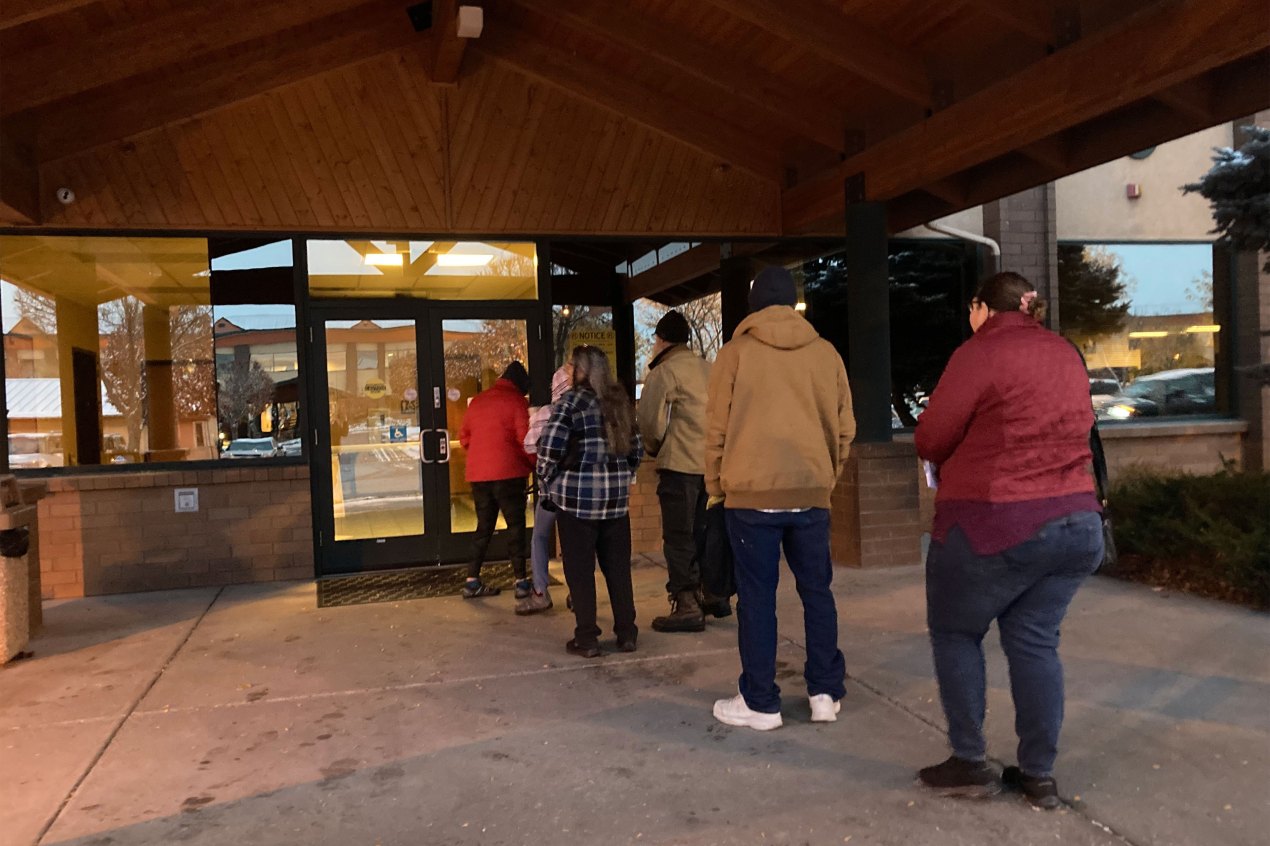 A line of six people are lined up outside the entrance doors of a building.