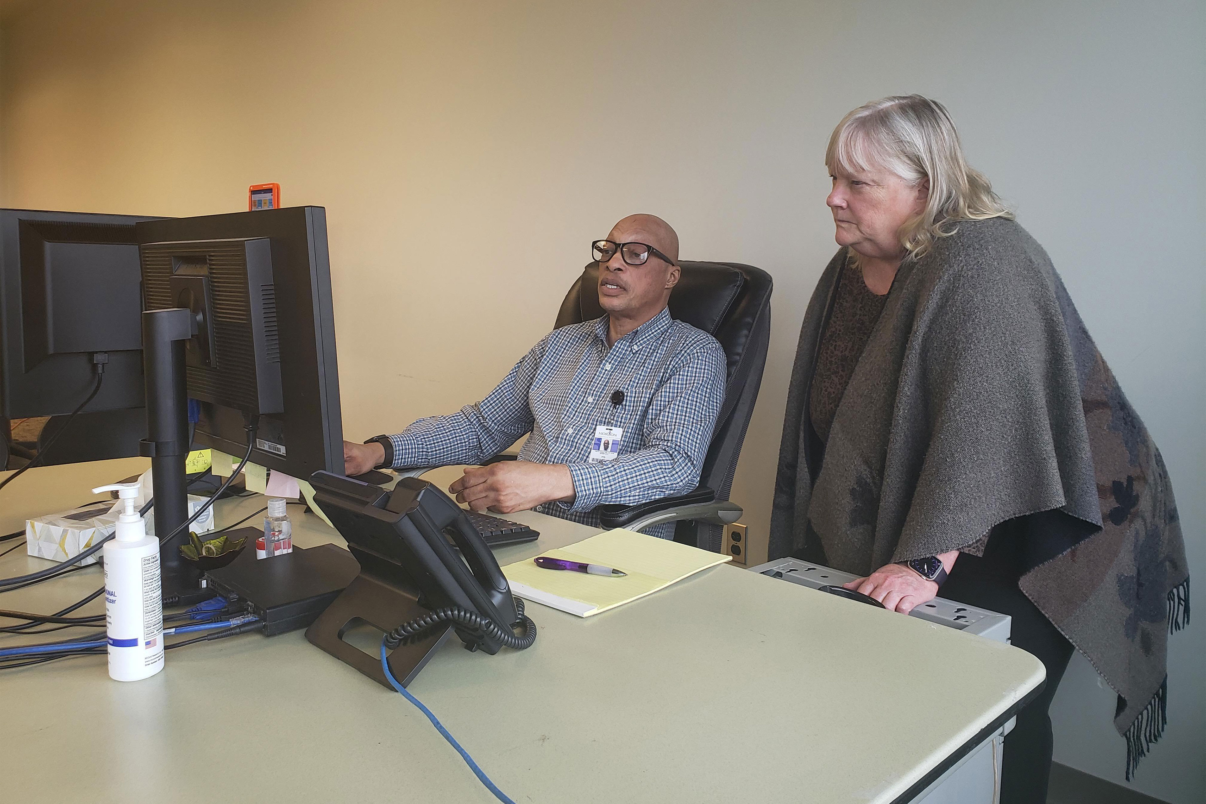 A man sits at a computer on a desk and a woman stands next to him and looks at the computer screen