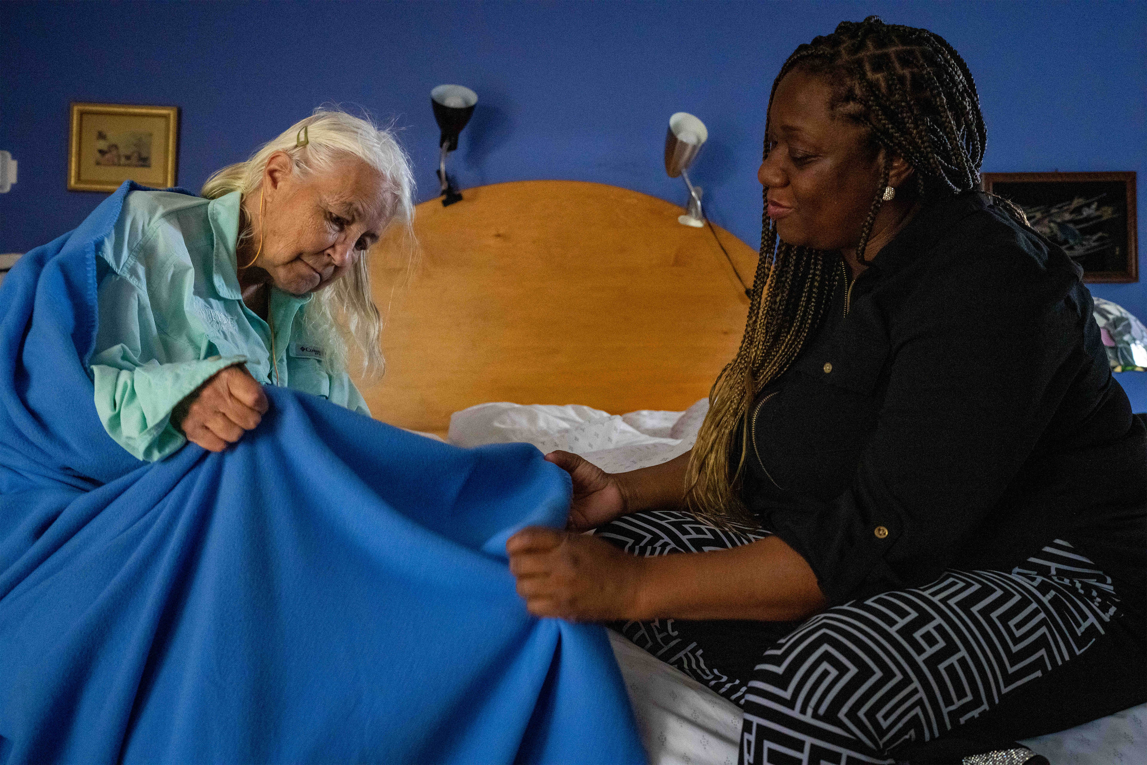A photo of a caregiver helping an elderly woman with a blanket in bed.