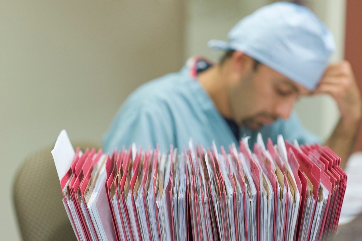 A health care worker rests their hand on their forehead behind stacks of paperwork.