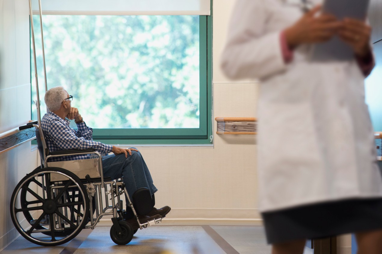 An elderly man is sitting in a wheelchair in a hospital room. A health care worker is visible in the foreground of the image.