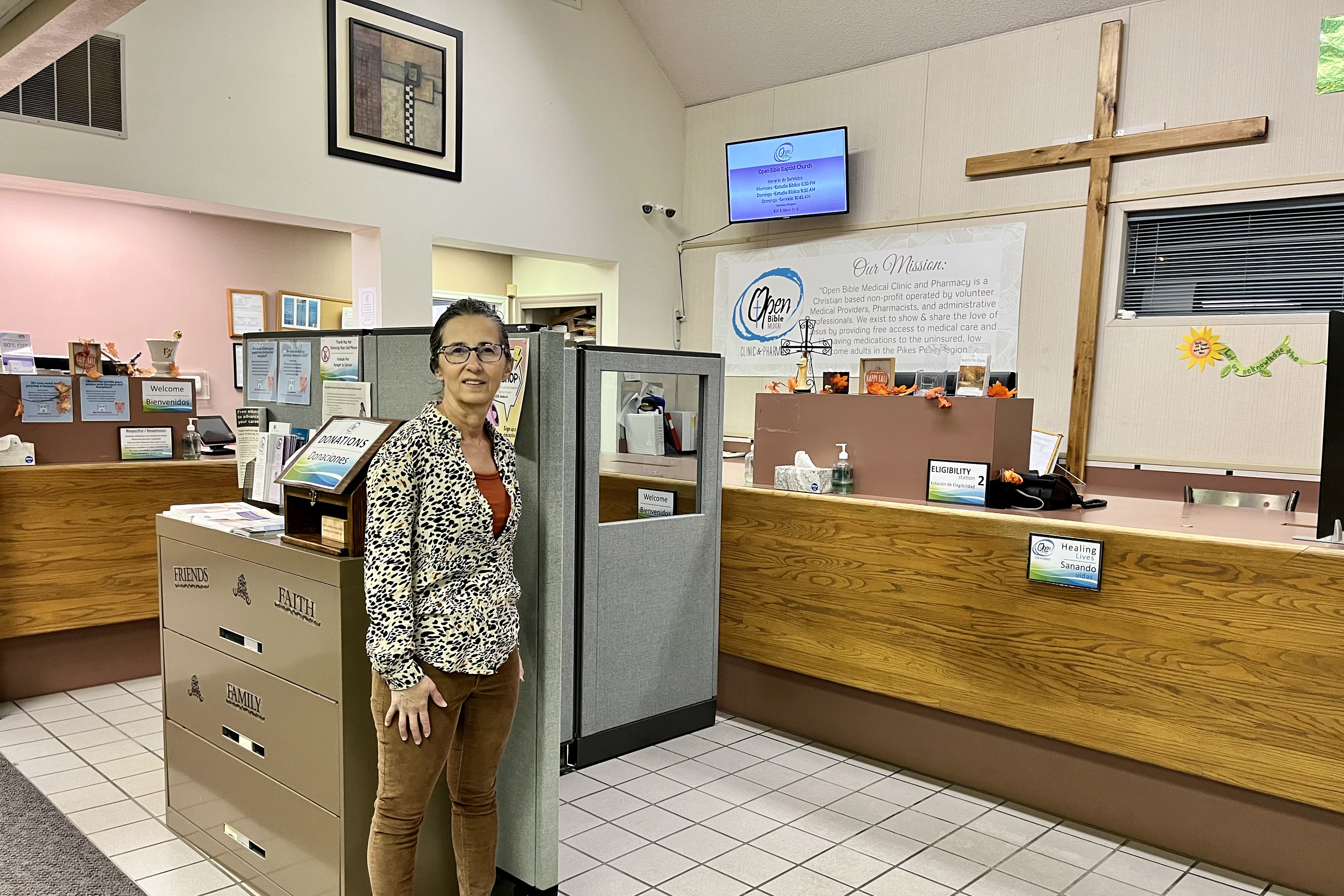 Pharmacist Frieda Martin poses at Open Bible Clinic and Pharmacy in Colorado Springs, Colorado, on Nov. 7, 2023. A large cross is on the wall behind her, along with signs and other information about the clinic.
