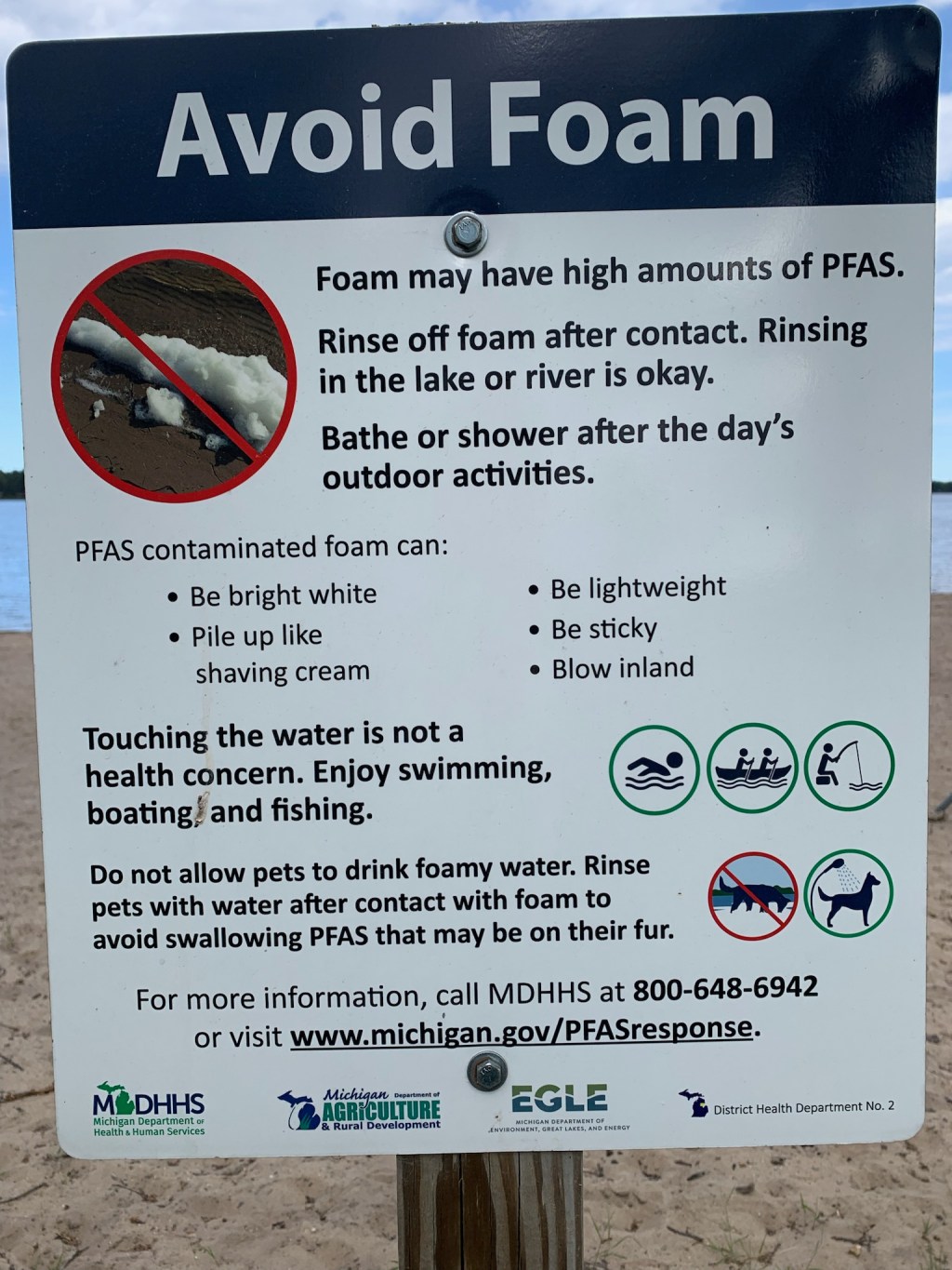 A sign is posted at a Van Etten Lake public beach in Oscoda, Michigan. It reads, "Avoid Foam / Foam may have high amounts of PFAS. Rinse off foam after contact. Rinsing in the lake or river is okay. Bathe or shower after the day's outdoor activities. / PFAS contaminated foam can be: Bright white; Pile up like shaving cream; Be lightweight; Be sticky; Blow inland / Touching the water is not a health concern. Enjoy swimming, boating, and fishing. / Do not allow pets to drink foamy water. Rinse pets with water after contact with foam to void swallowing PFAS that may be on their fur." It includes contact information at the bottom of the the sign.