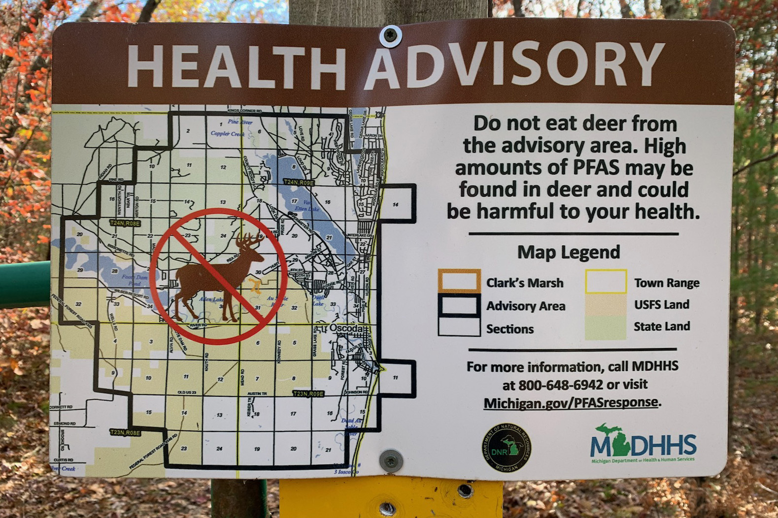 A health advisory sign posted at Clark’s Marsh reads, "HEALTH ADVISORY / Do not eat deer from the advisory area. High amounts of PFAS may be found in deer and could be harmful to your health." The sign includes a map and a legend that further details the area.