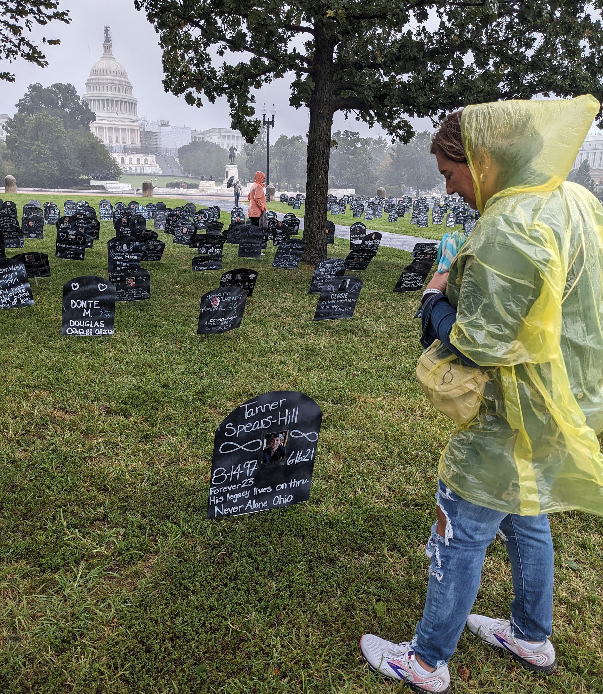 A woman wearing a transparent yellow rain jacket looks down at a sign stuck into the ground. There are dozens of similar markers in the background and the U.S. Capitol farther in the distance.
