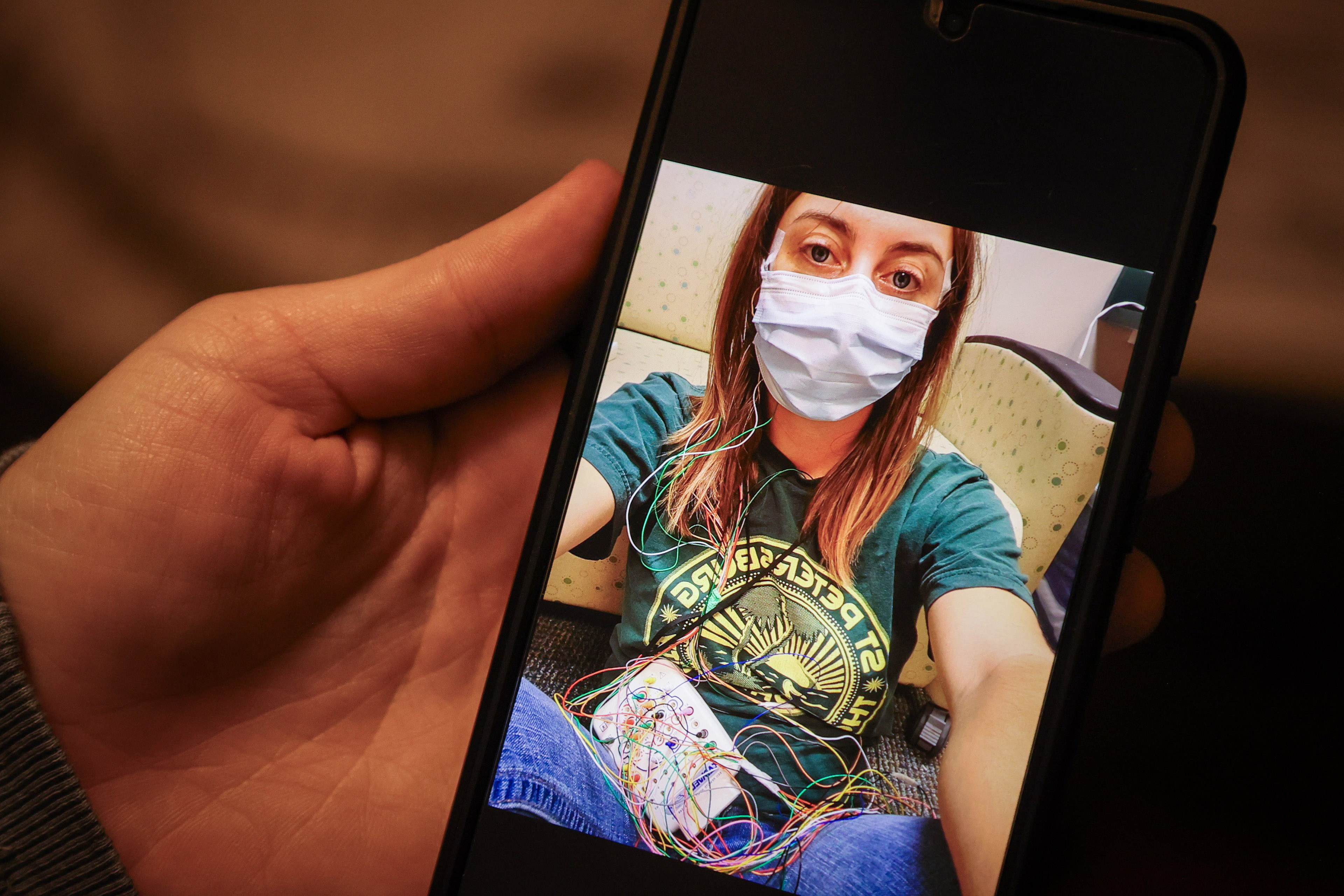 Nina Shand shows a photo of herself on her phone from when she was in a sleep study testing for narcolepsy. In the photo, she wears a surgical mask and is collected to many thin, colorful wires.